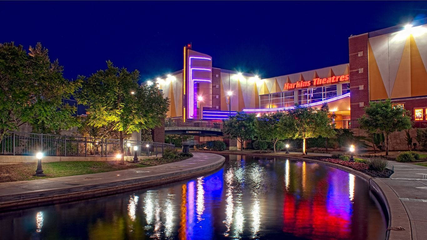 德州 桌布 壁紙 — 現代劇院建築的充滿活力的夜景，霓虹燈在彎曲的水道上反射，周圍環繞著樹木和小徑。