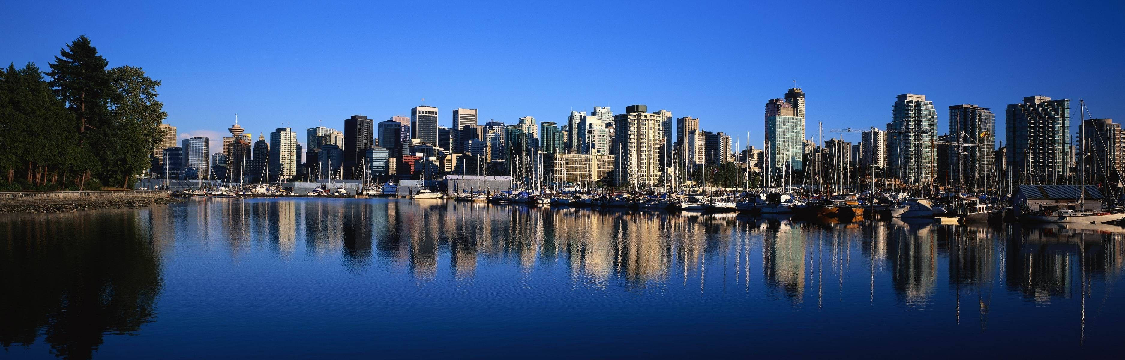 藍天 桌布 壁紙 — 現代城市天際線的全景視圖，完美倒映在平靜的藍色水面上，前景有停泊的帆船。
