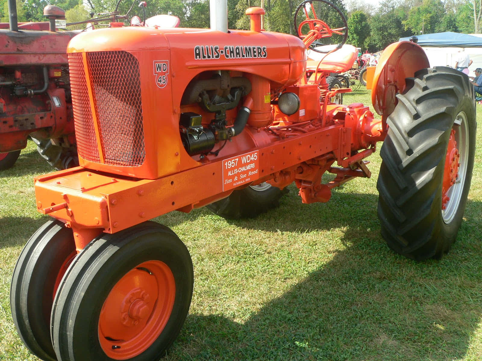 現代強勁拖拉機在田間耕作 壁紙 — 鮮豔的橙色 1957 年 Allis-Chalmers WD45 拖拉機停在草地上，背景有樹木，陽光普照。