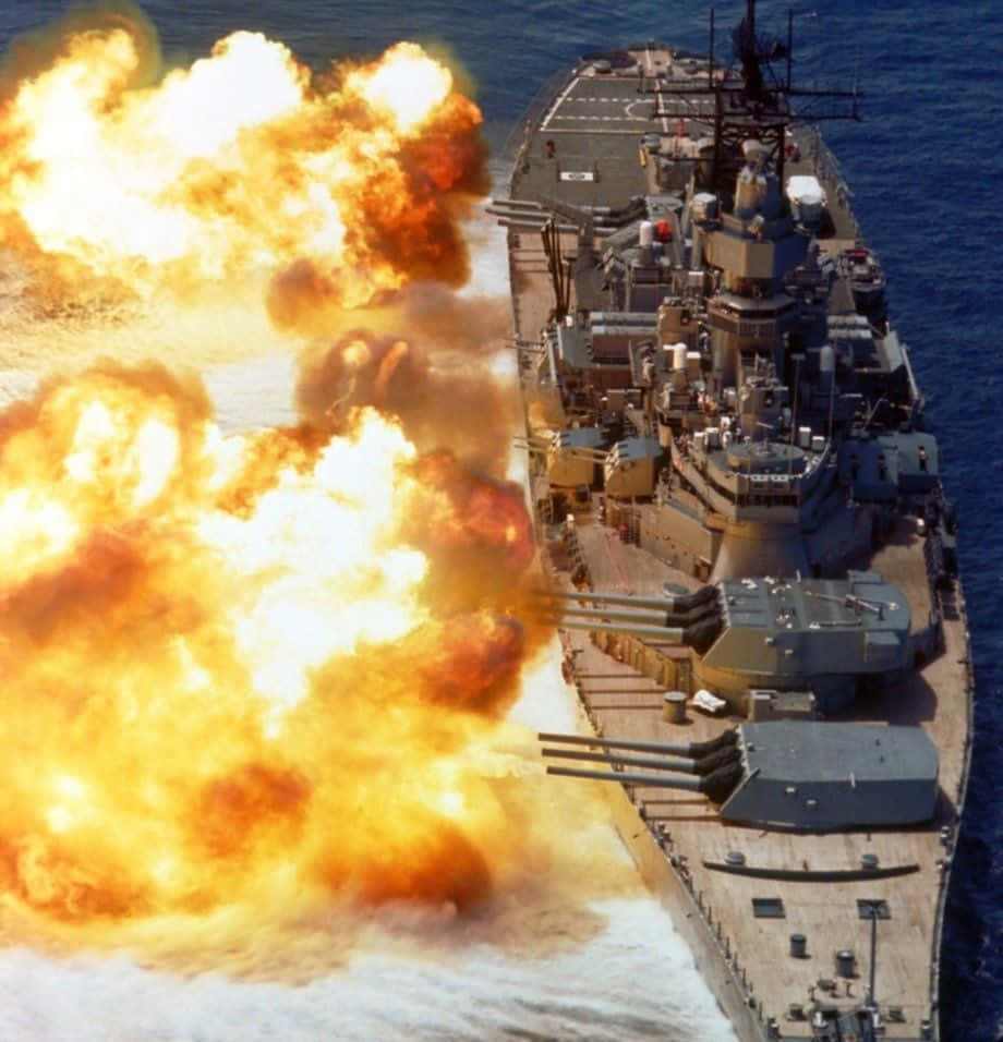 現代戰艦全速航行 壁紙 — 一艘戰艦發射巨型火炮，在晴朗的藍天下向海洋中噴射出熾熱的橙色爆炸。