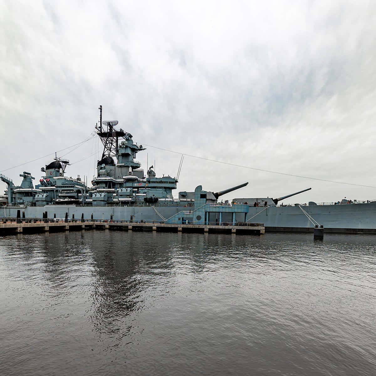 現代戰艦與驅逐艦巡航於開闊海域 壁紙 — 一艘巨大的灰色戰艦停泊在碼頭，天空多雲，平靜的水面映照出其宏偉的結構。