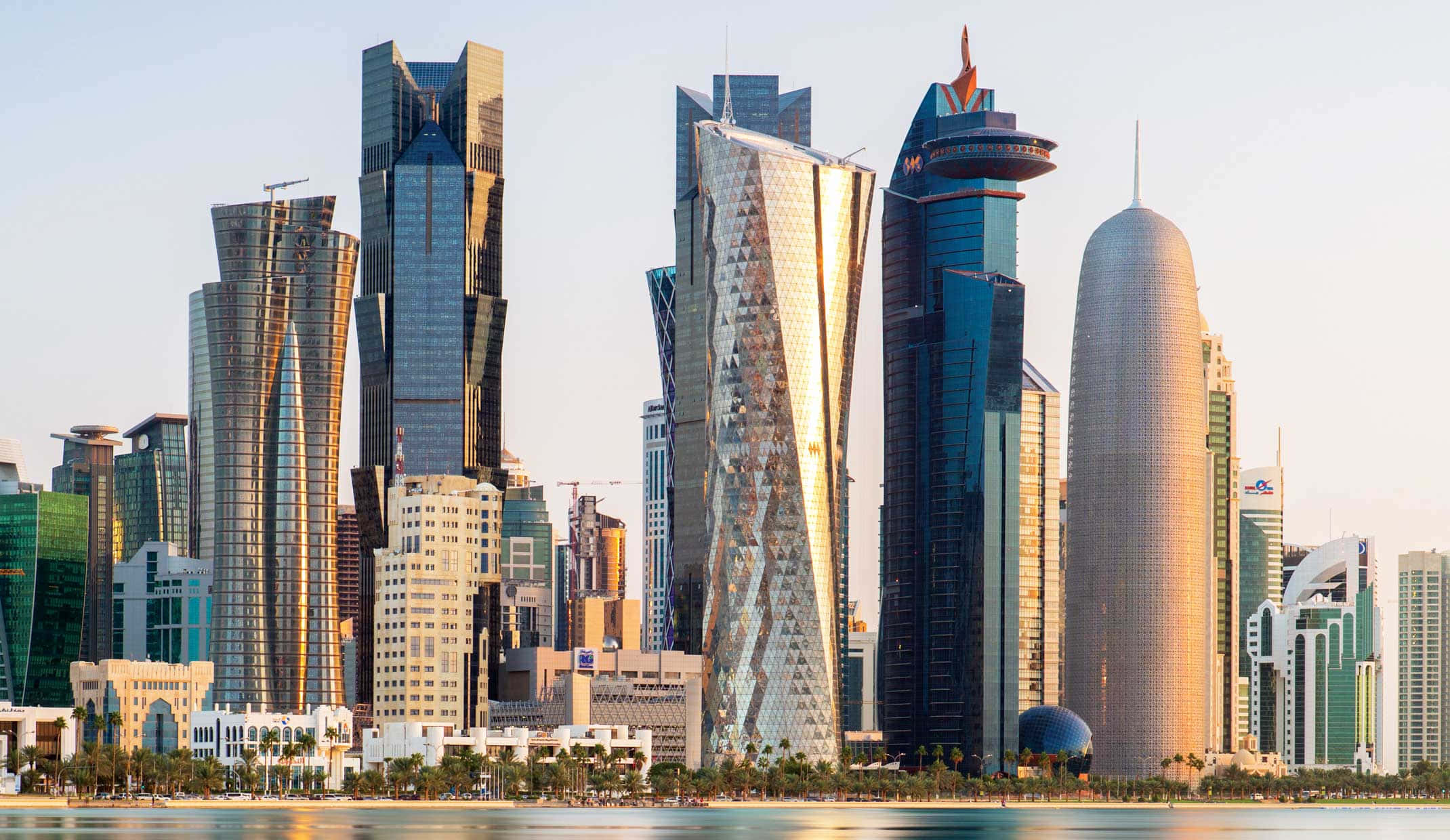 Qatar 桌布 壁紙 — 現代摩天大樓以反光玻璃幕牆在平靜水面上柔和金色夕陽天空的襯托下戲劇性地聳立。