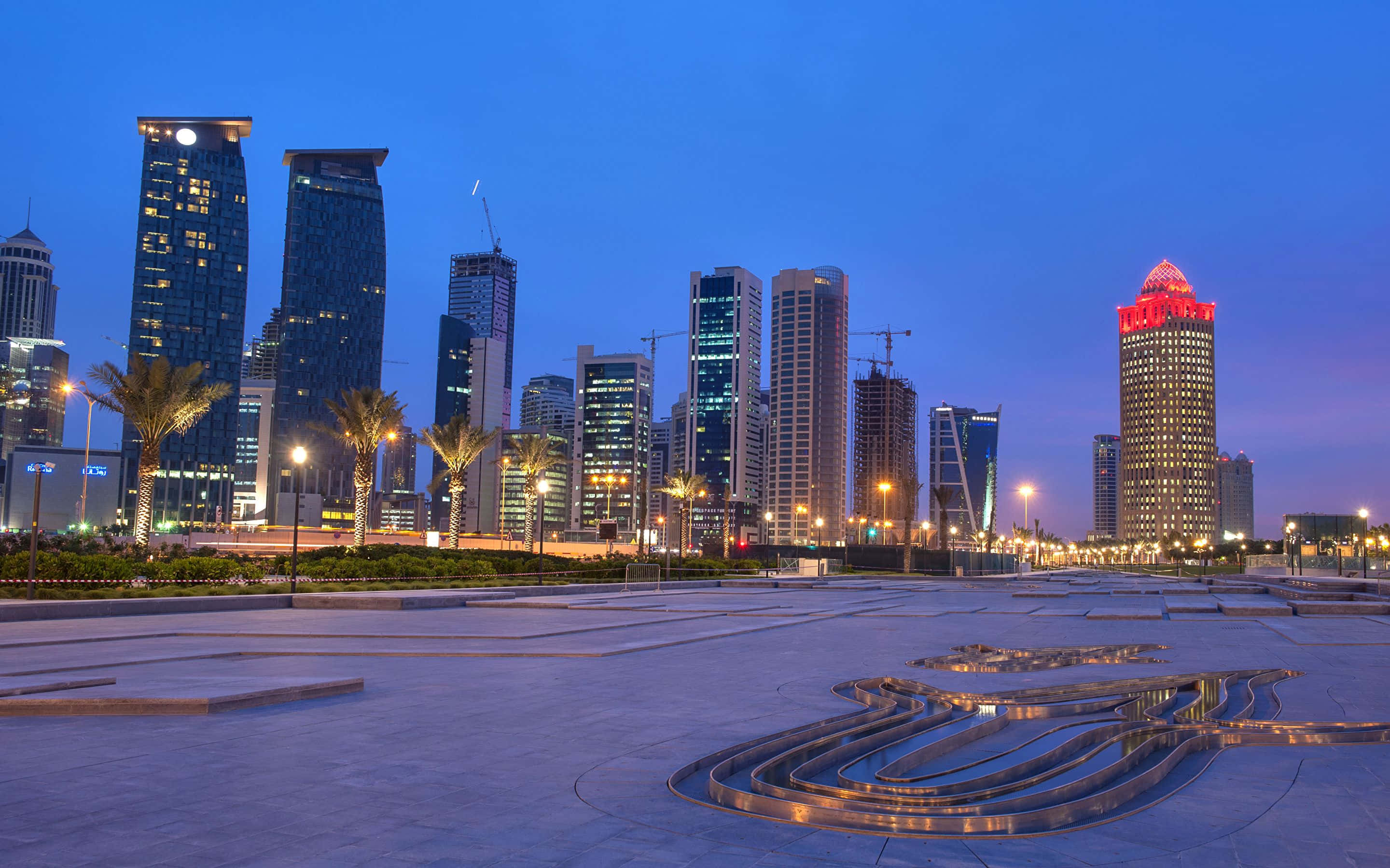 Qatar 桌布 壁紙 — 現代摩天大樓在暮色天空下閃耀，棕櫚樹排列在反射廣場兩旁，廣場上有彎曲的金屬水景。