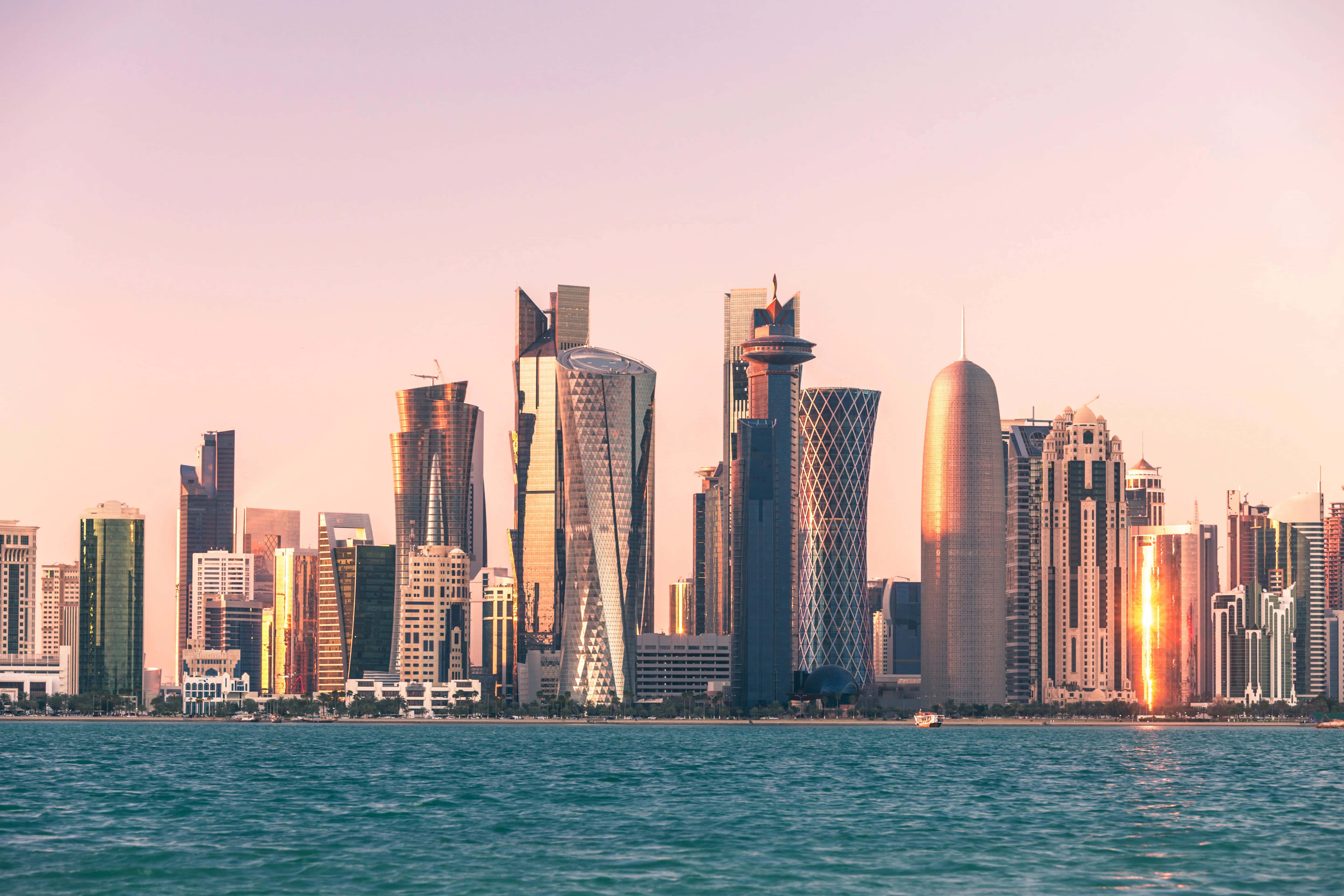 Qatar 桌布 壁紙 — 現代摩天大樓採用反光玻璃外牆，在海濱於黃金時刻升起，在碧綠的海面上投射出溫暖的光芒。