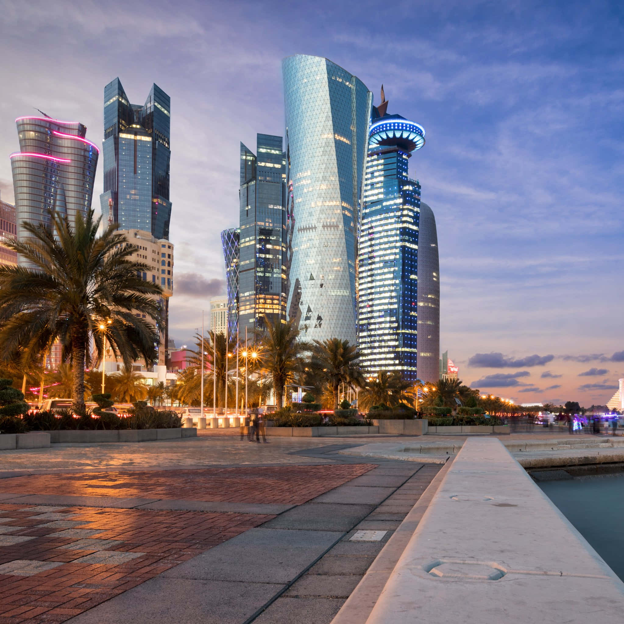 Qatar 桌布 壁紙 — 現代摩天大樓散發著藍色和粉紅色的光芒，在黃昏時分沿著海濱長廊聳立在棕櫚樹之上。