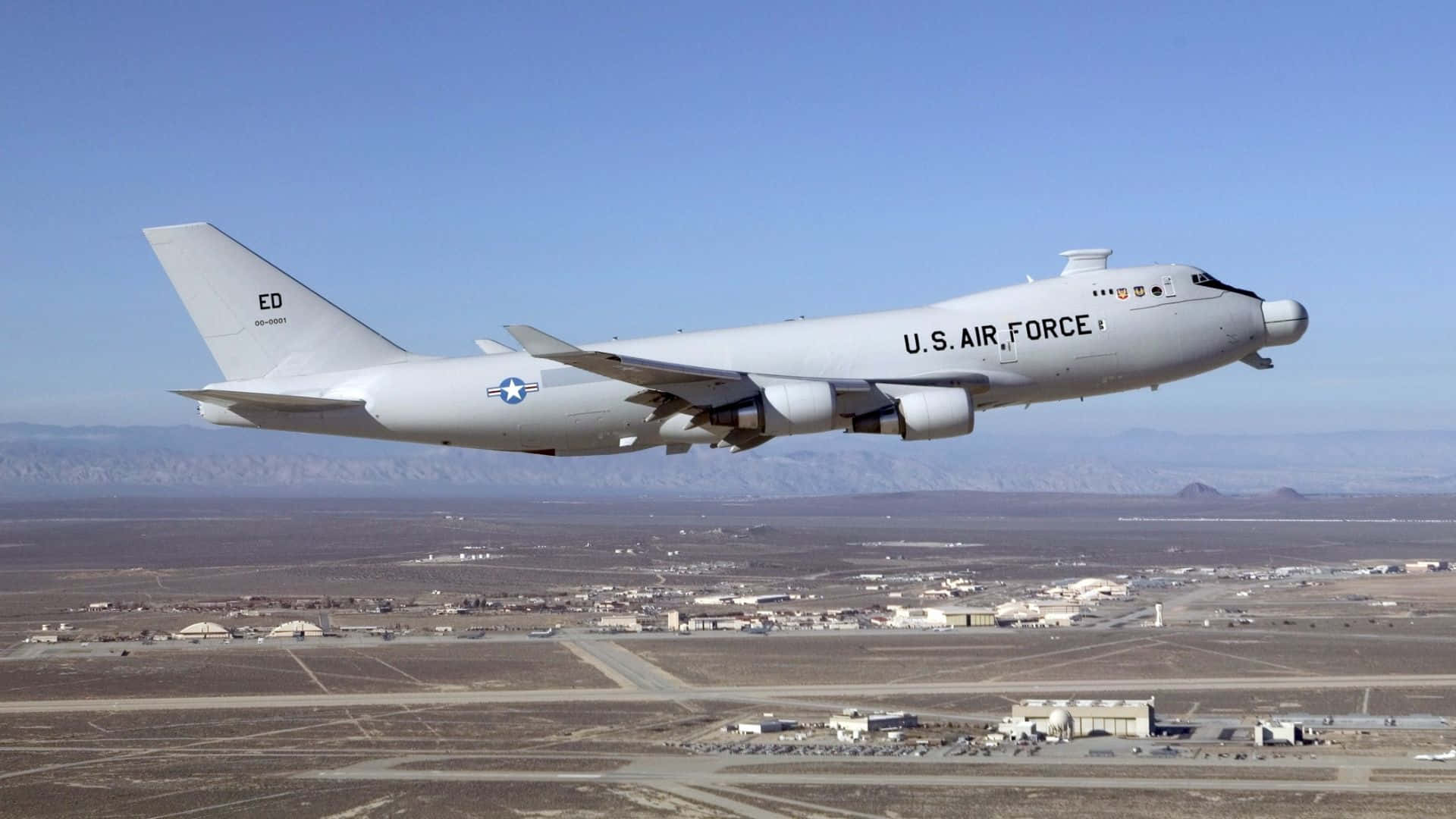 現代時尚大型客機騰空而起 壁紙 — 一架美國空軍KC-135同溫層加油機在晴朗藍天下，飛越沙漠空軍基地，遠方山巒起伏。