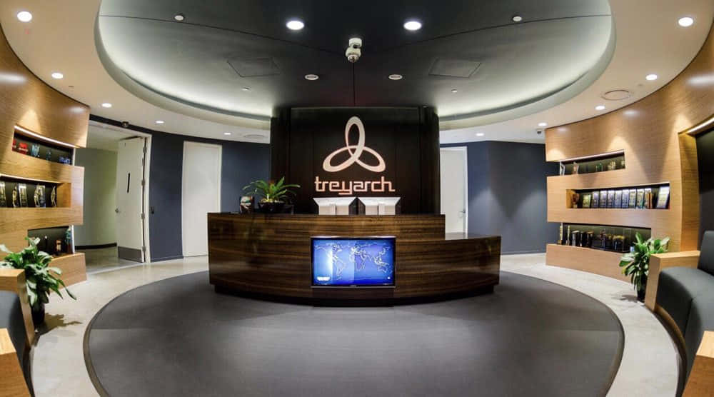Treyarch 桌布 壁紙 — 現代時尚的企業大堂，設有彎曲木製接待處，展示 Treyarch 標誌和嵌入式屏幕，周圍環繞著溫暖的木架和盆栽植物。