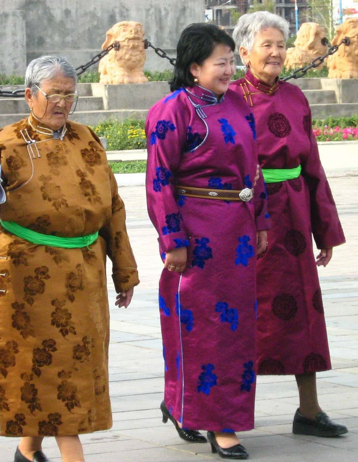 現代服裝蒙古女性畫面 壁紙 — 三位身穿傳統刺繡長袍的老年女性一起走在鋪砌的廣場上，背景有石獅雕像。
