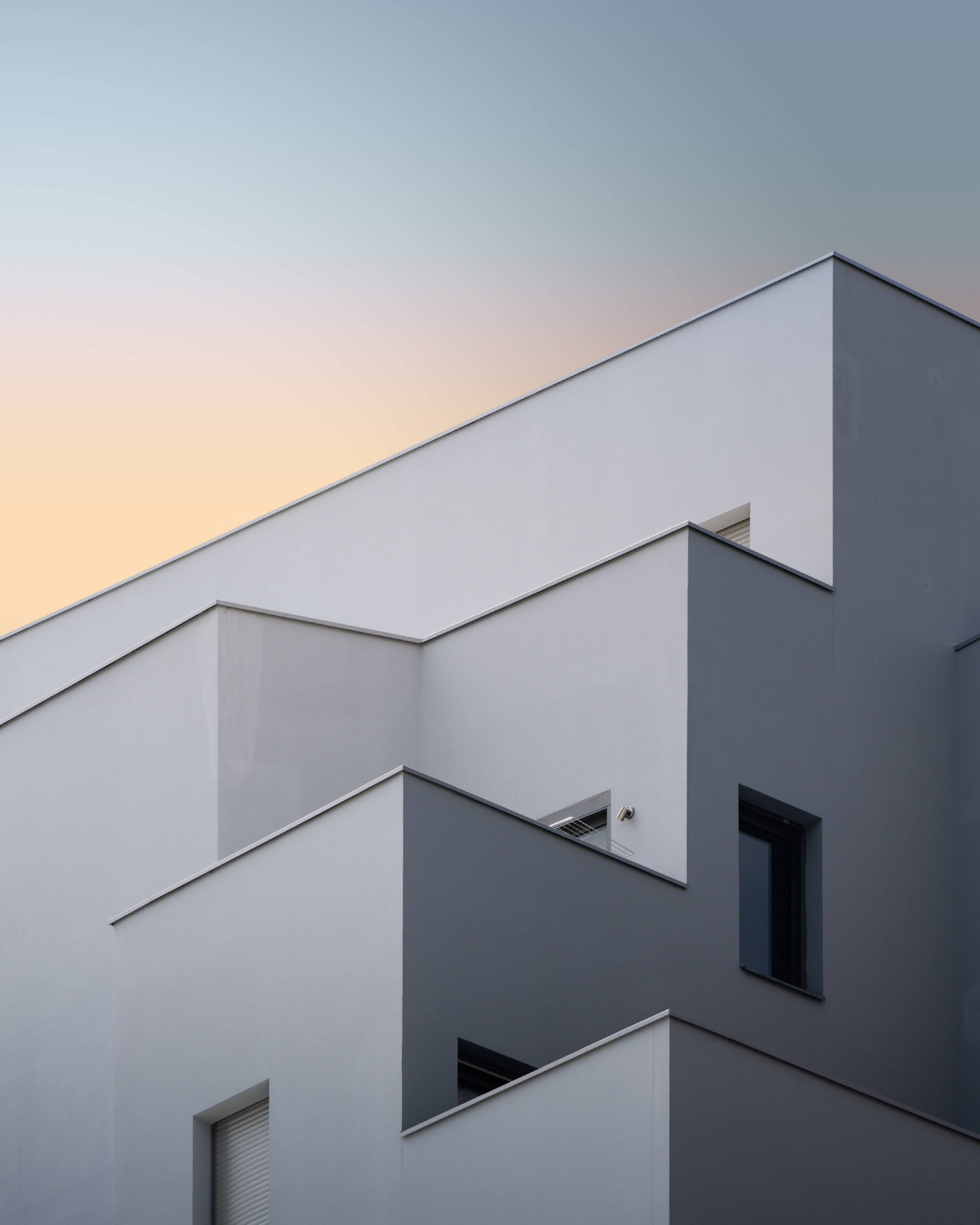 現代極簡主義建築 壁紙 — 現代幾何建築外牆，以純淨的白色和灰色平面襯托柔和的日落天空。