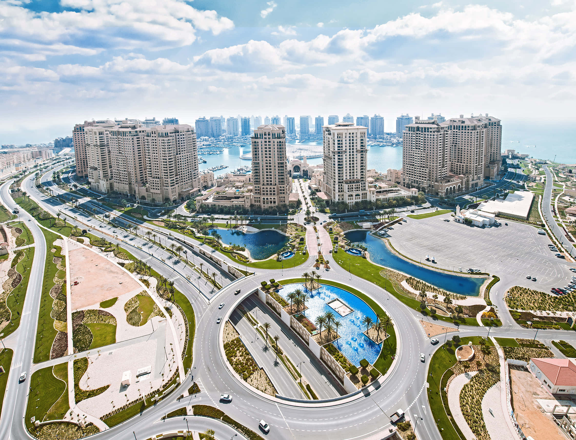 Qatar 桌布 壁紙 — 現代沿海城市的鳥瞰圖，彎曲的高速公路蜿蜒穿過園林公園和高層建築，在明亮的藍天和蓬鬆的白雲下。