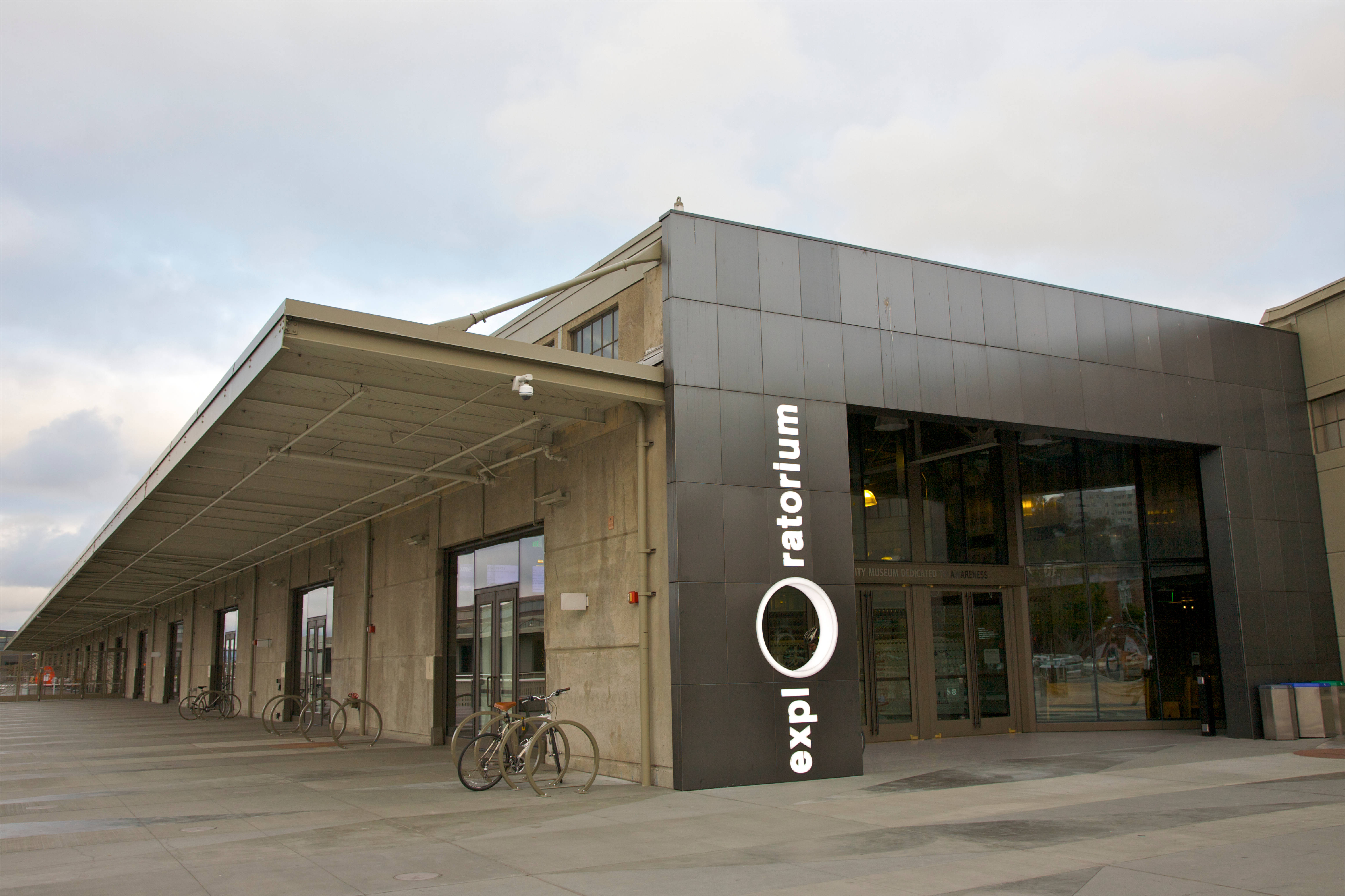 三藩市 桌布 壁紙 — 現代混凝土和玻璃建築，帶有 'exploratorium' 標誌，單車停放在外面，天空多雲。