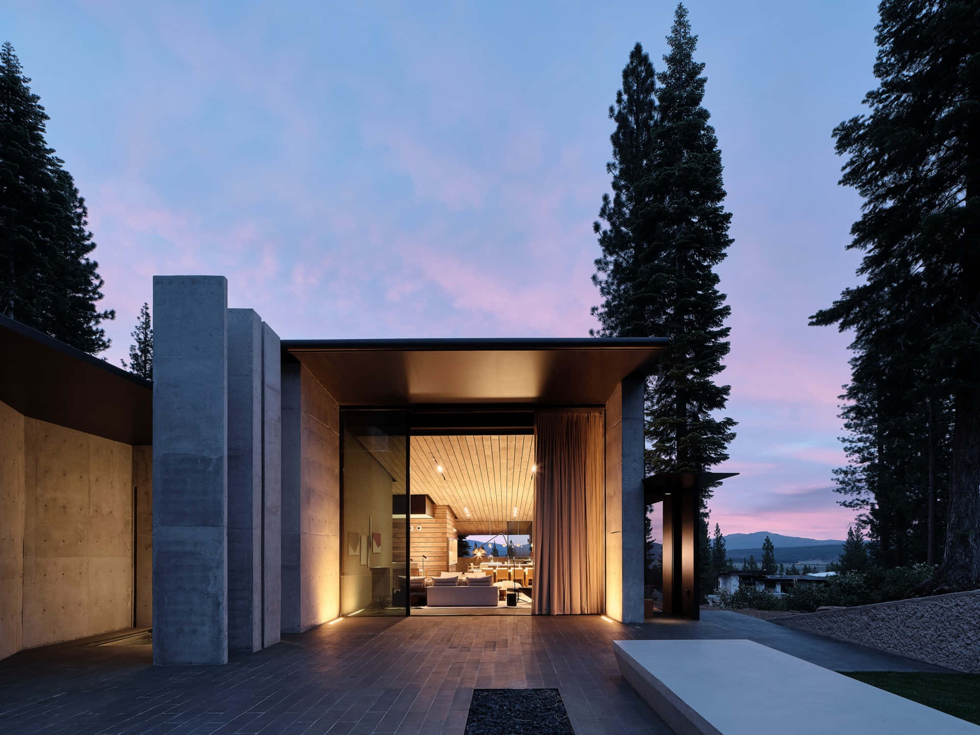 Minimalist Architecture 桌布 壁紙 — 現代混凝土房屋敞開，露出溫暖的室內光芒，映襯著暮色天空和高大的松樹。