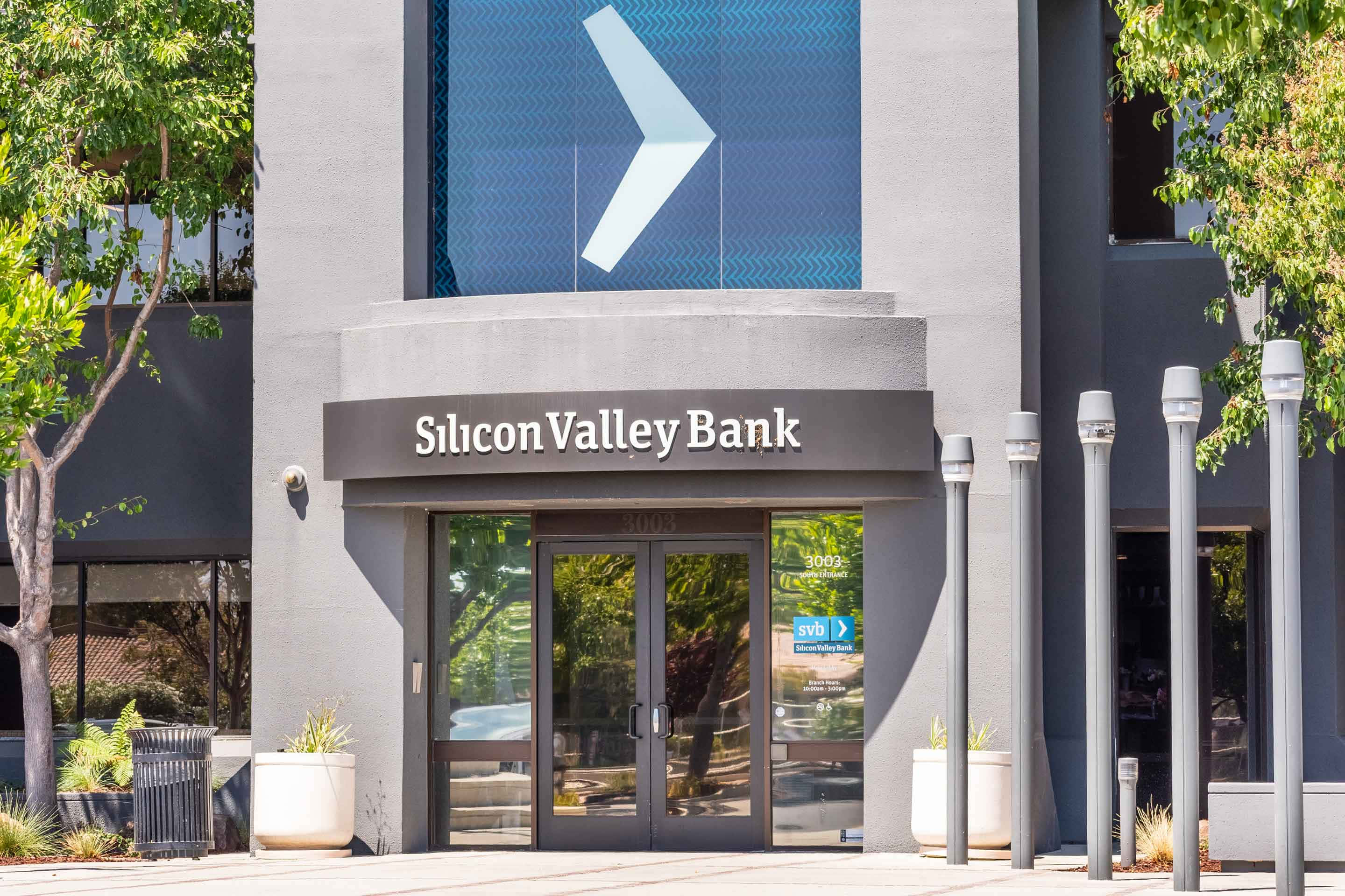 Bank 桌布 壁紙 — 現代灰色矽谷銀行大樓，設有玻璃門和藍色V形標誌，在明亮的日光下。