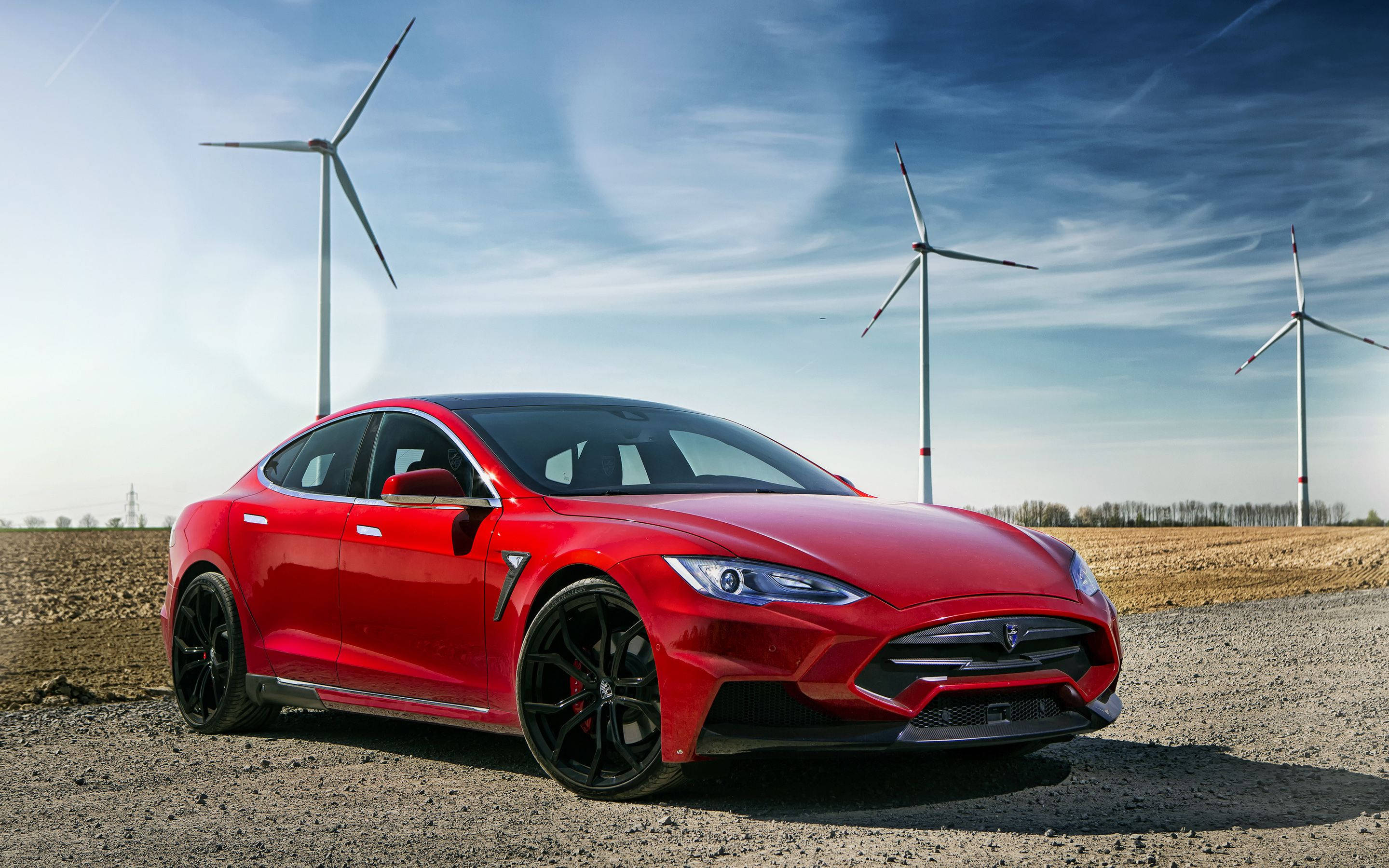 現代特斯拉 Model S 與 Larte 設計 壁紙 — 一輛亮紅色的Tesla Model S Performance停泊在碎石上，三座風力發電機高聳在明亮的藍天下。