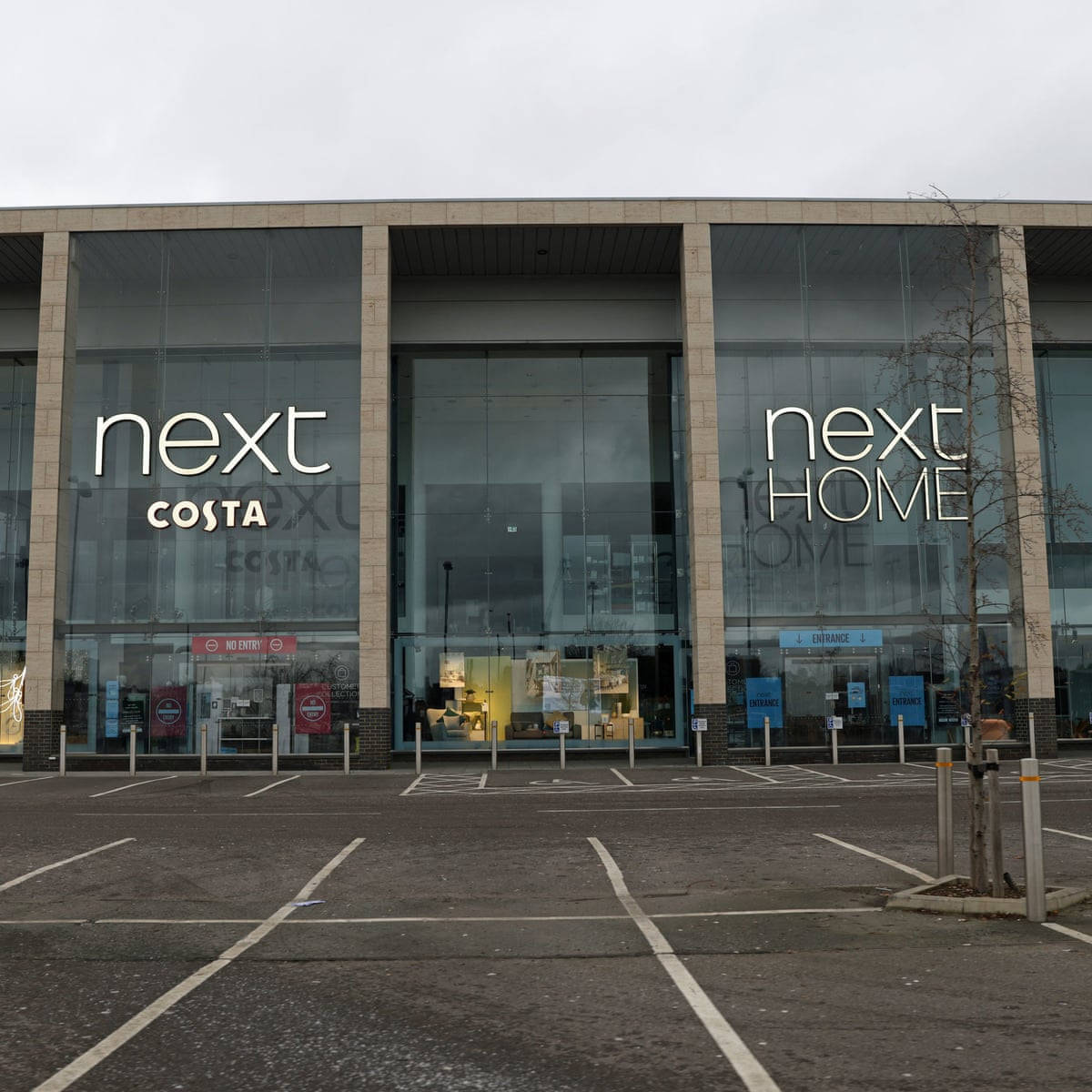Next Home 壁紙 — 現代玻璃幕牆的Next和Costa商店，天空多雲，停車場空無一人，樹木光禿。