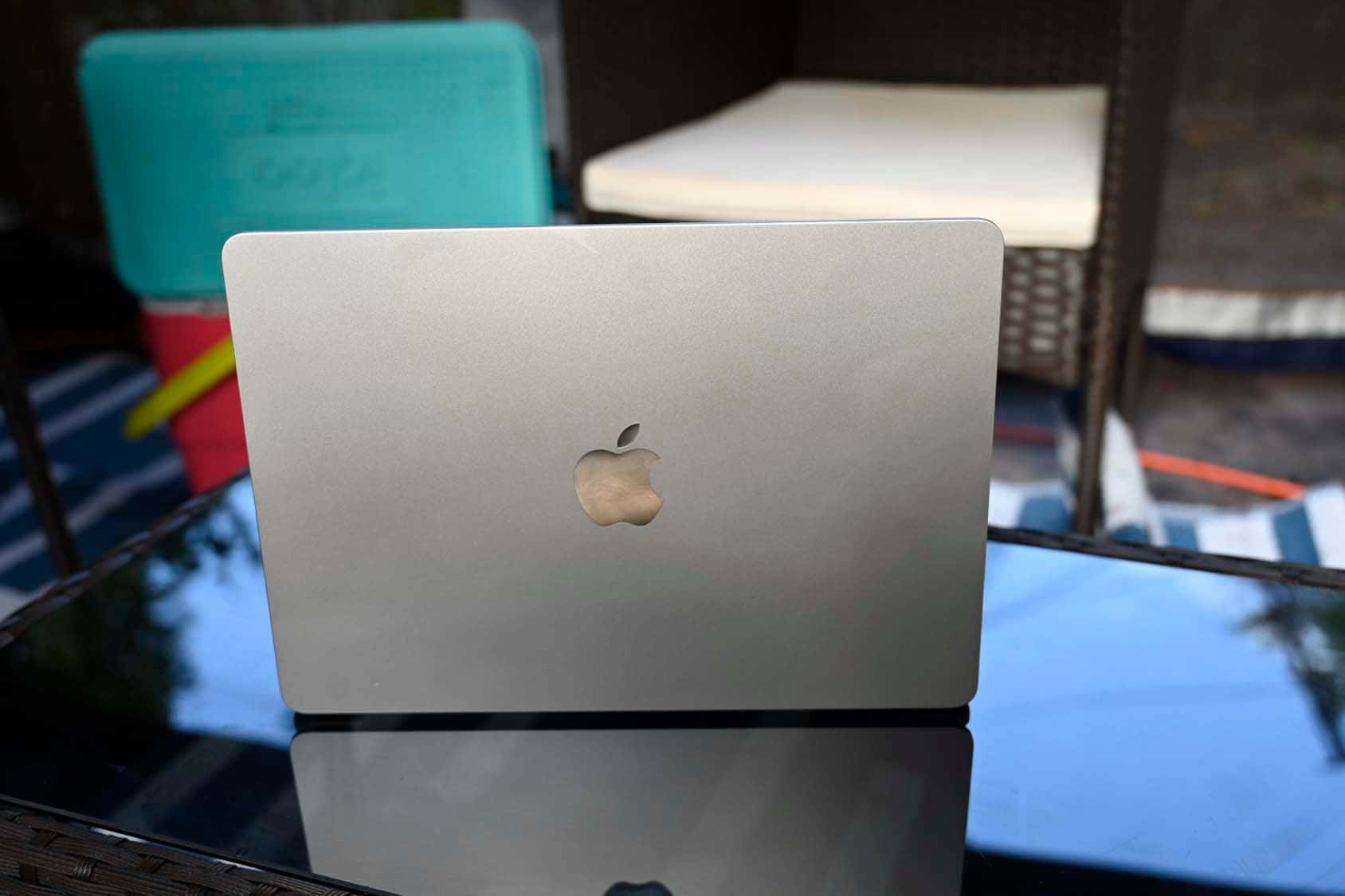 現代筆電與咖啡擺放在木質書桌 壁紙 — 銀色MacBook蓋上帶有蘋果標誌，放在玻璃桌上，反射藍天和戶外庭院家具。