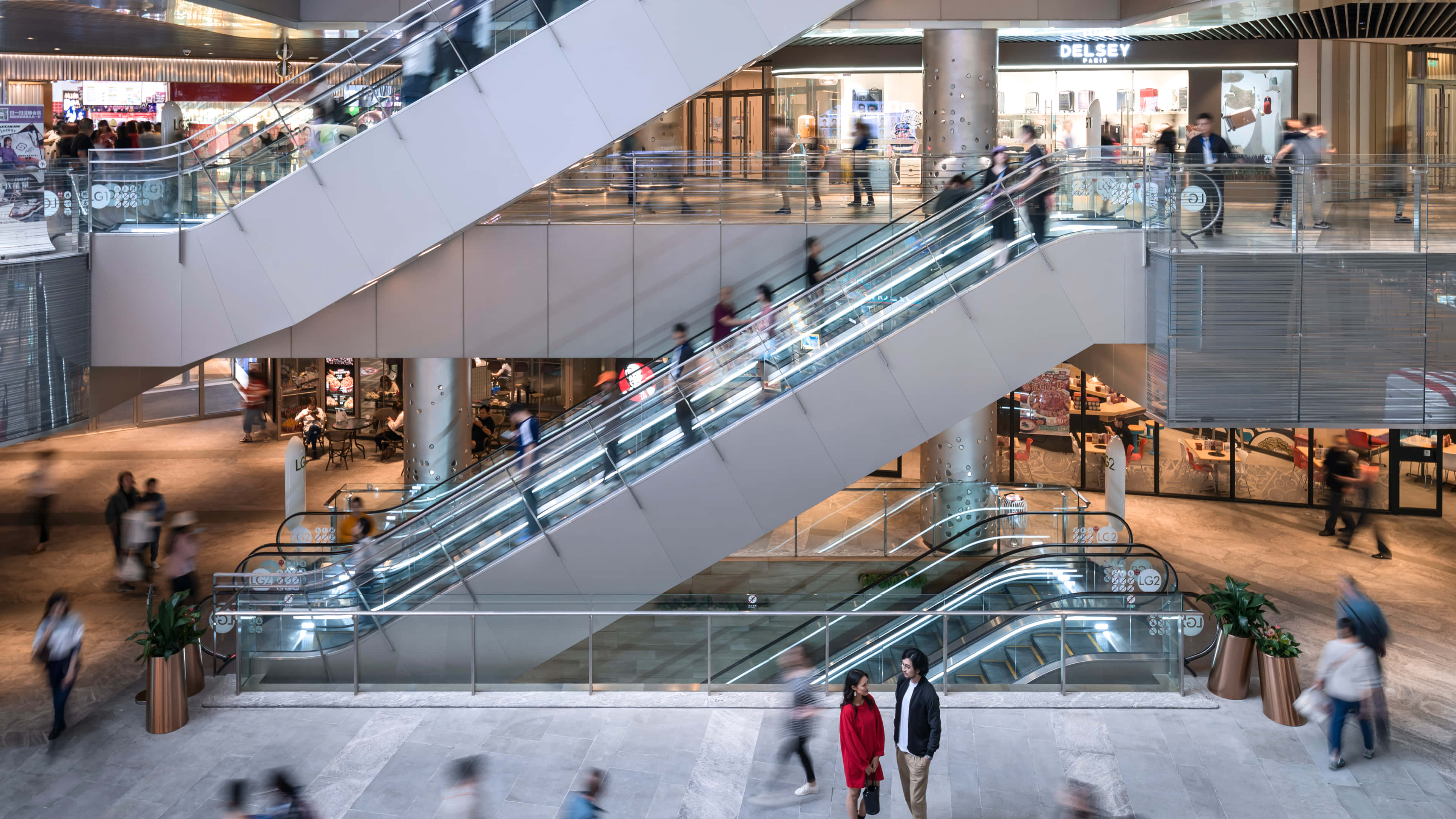Mall 桌布 壁紙 — 現代購物中心，多條自動扶梯在明亮的人造燈光下運送模糊的購物者穿梭於各樓層之間。