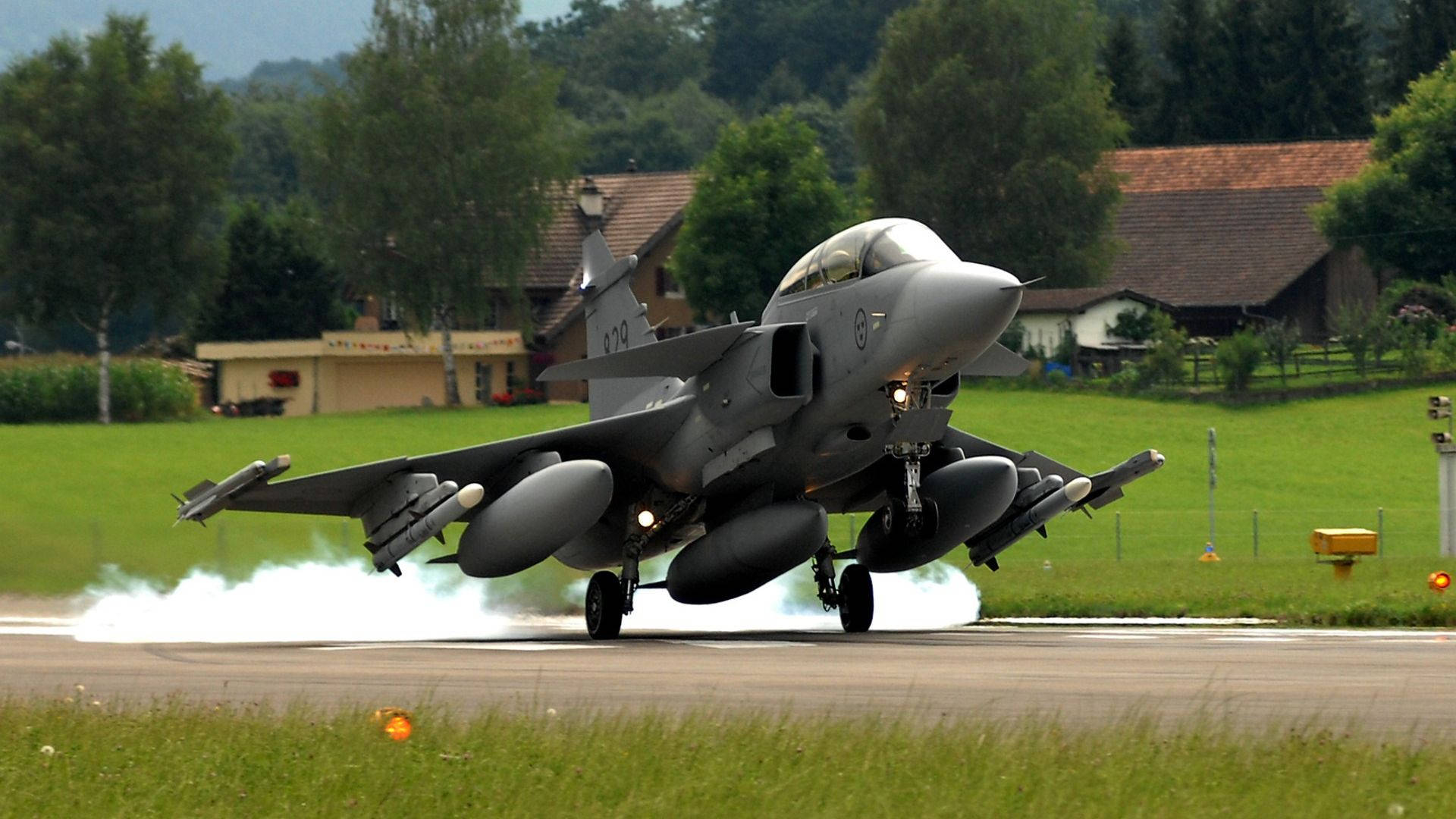 現代軍用戰鬥機翱翔天際 壁紙 — 一架灰色戰鬥機從跑道起飛，拖著煙霧，升越綠色田野和房屋。