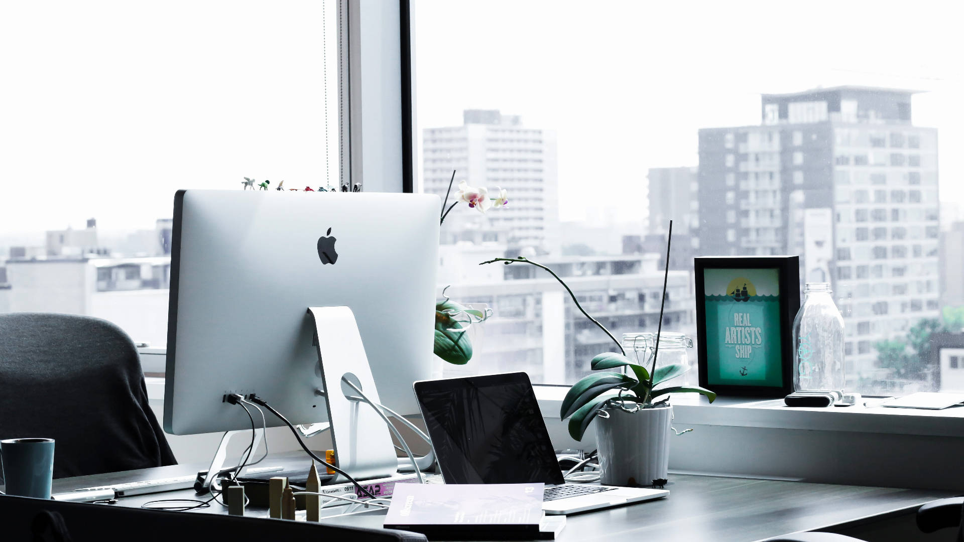 電腦 桌布 壁紙 — 現代辦公室書桌，配備 iMac 和手提電腦，位於窗邊，俯瞰城市天際線，並有盆栽植物和裱框藝術品。