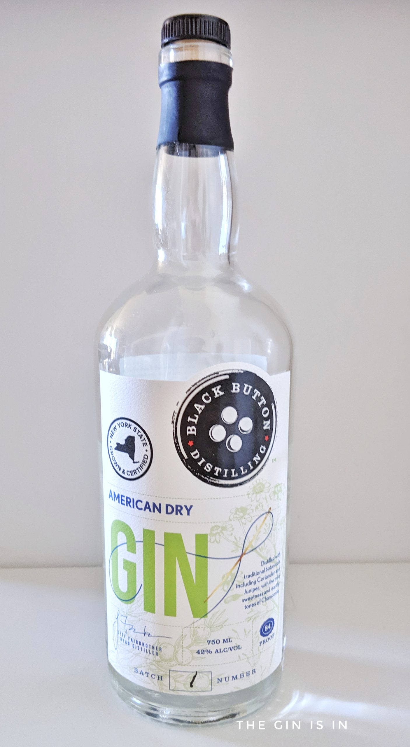 現代金酒黑按鈕蒸餾廠 壁紙 — 一個透明玻璃瓶的 Black Button Distilling American Dry Gin 直立於白色表面上，背景簡潔。