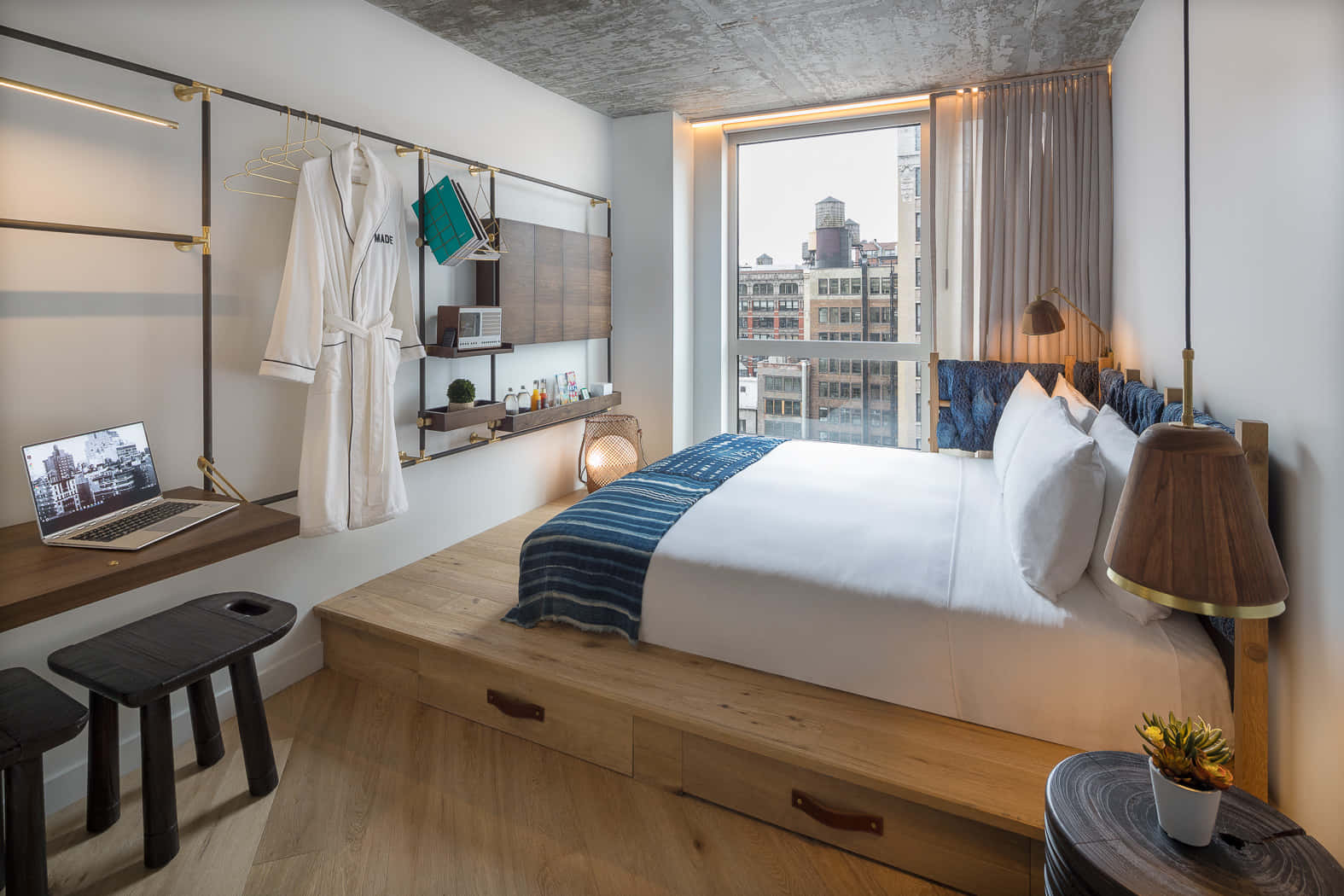 Hotel Room 桌布 壁紙 — 現代閣樓臥室，配有木製平台床、條紋藍色毯子，透過大窗戶可欣賞城市景觀。