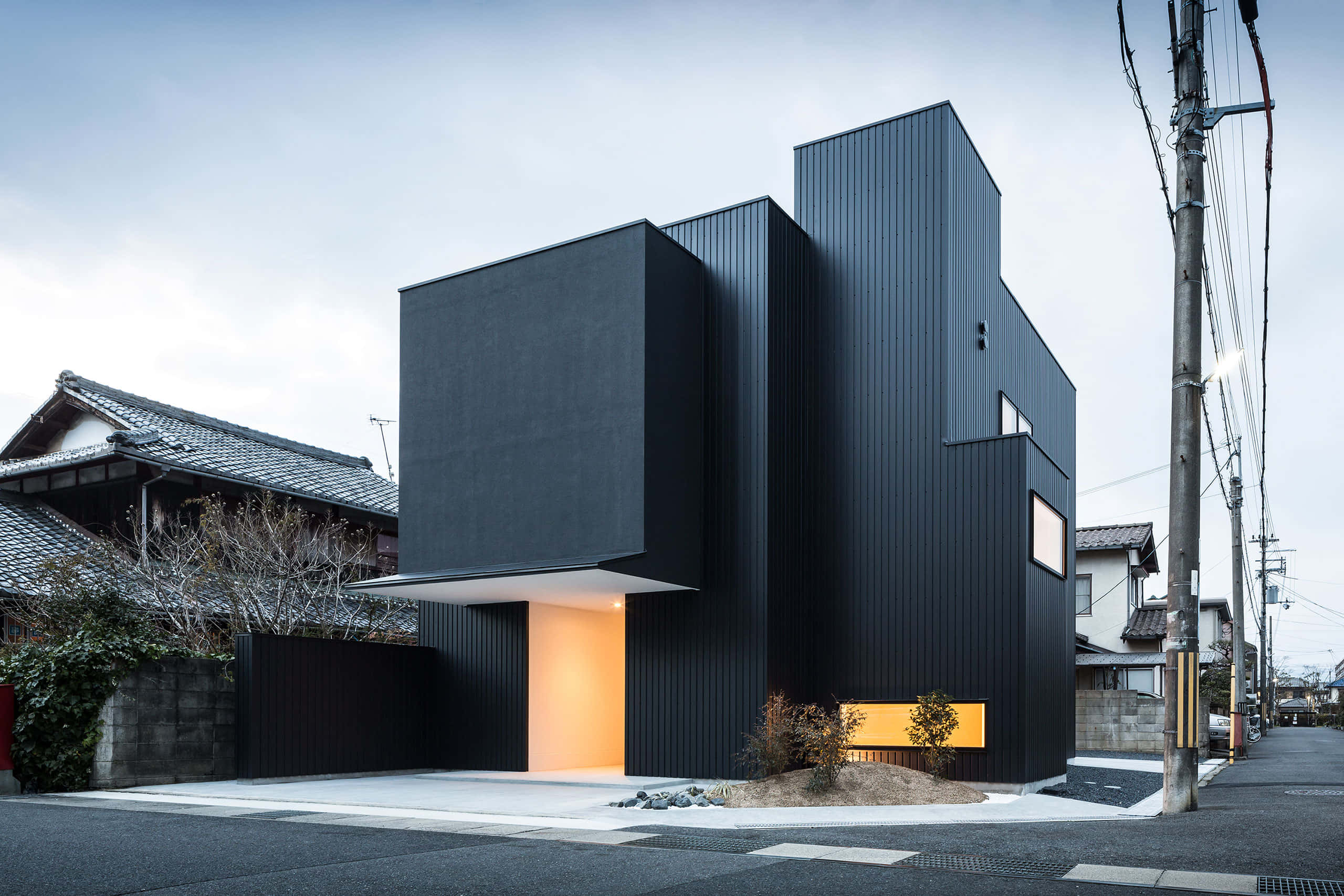 Minimalist Architecture 桌布 壁紙 — 現代黑色幾何房屋，發光的橙色入口與陰天下的傳統瓦屋頂形成對比。