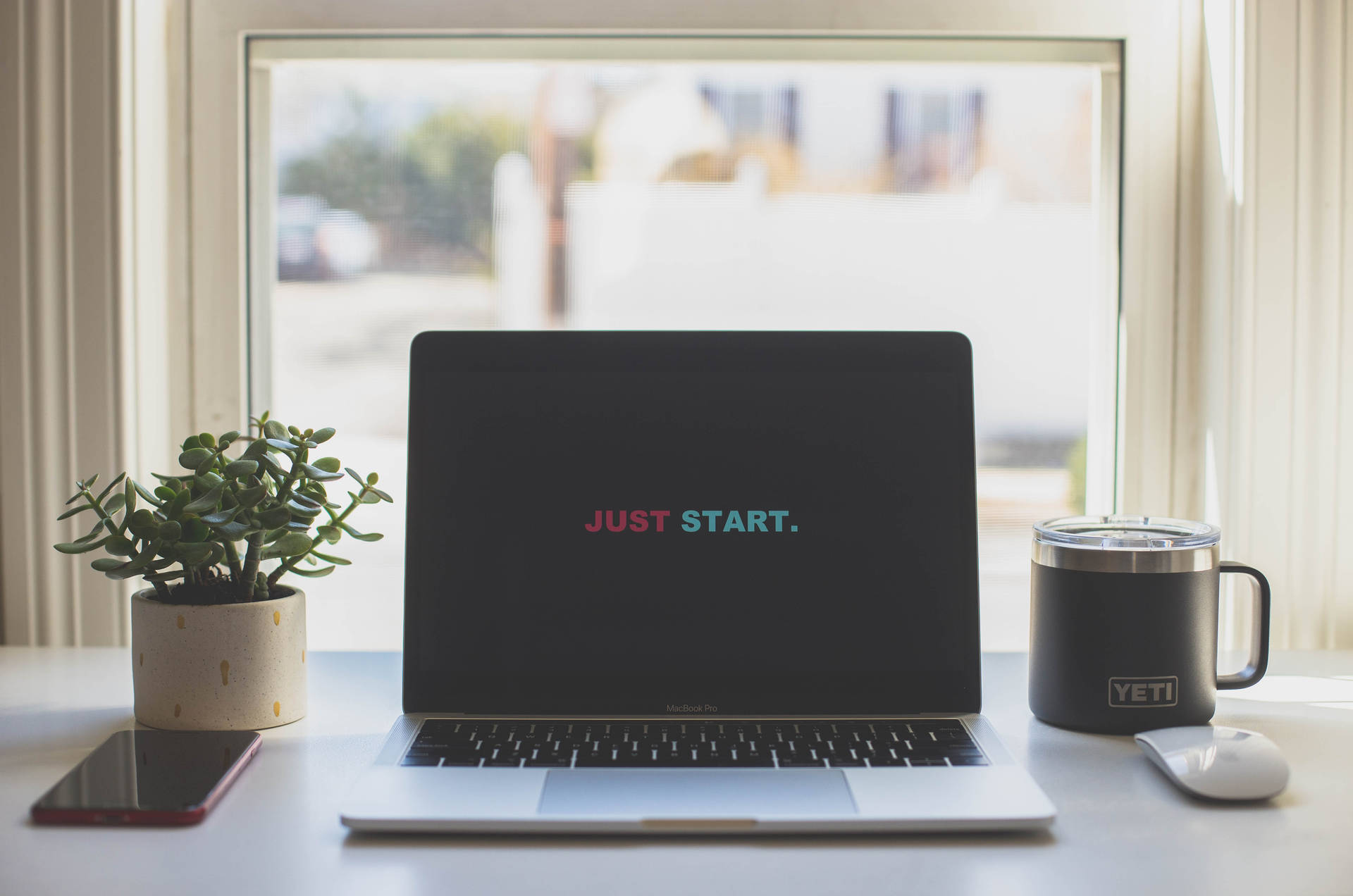 螢幕顯示「JUST START.」的筆記型電腦，旁邊有植物、杯子和電話在窗台上。
