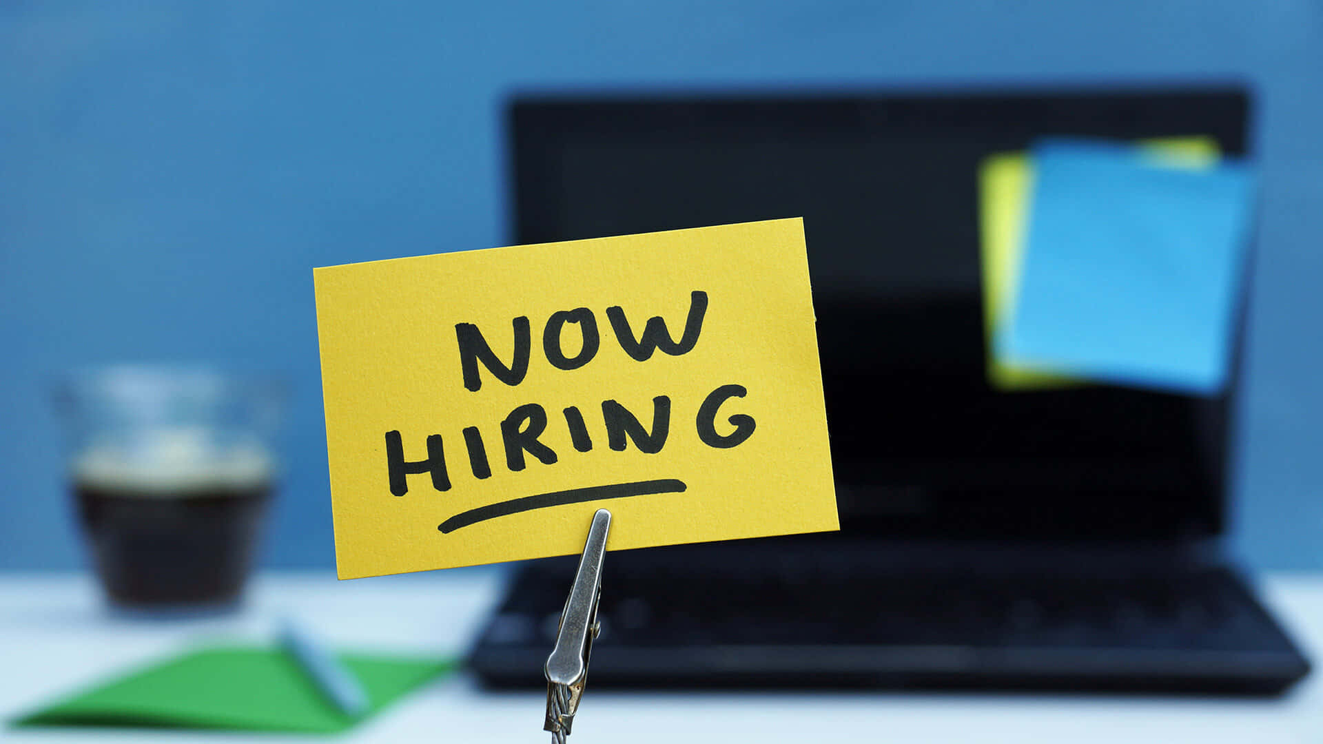 寫著 now hiring 的黃色便利貼夾在桌上的手提電腦上，旁邊有咖啡。
