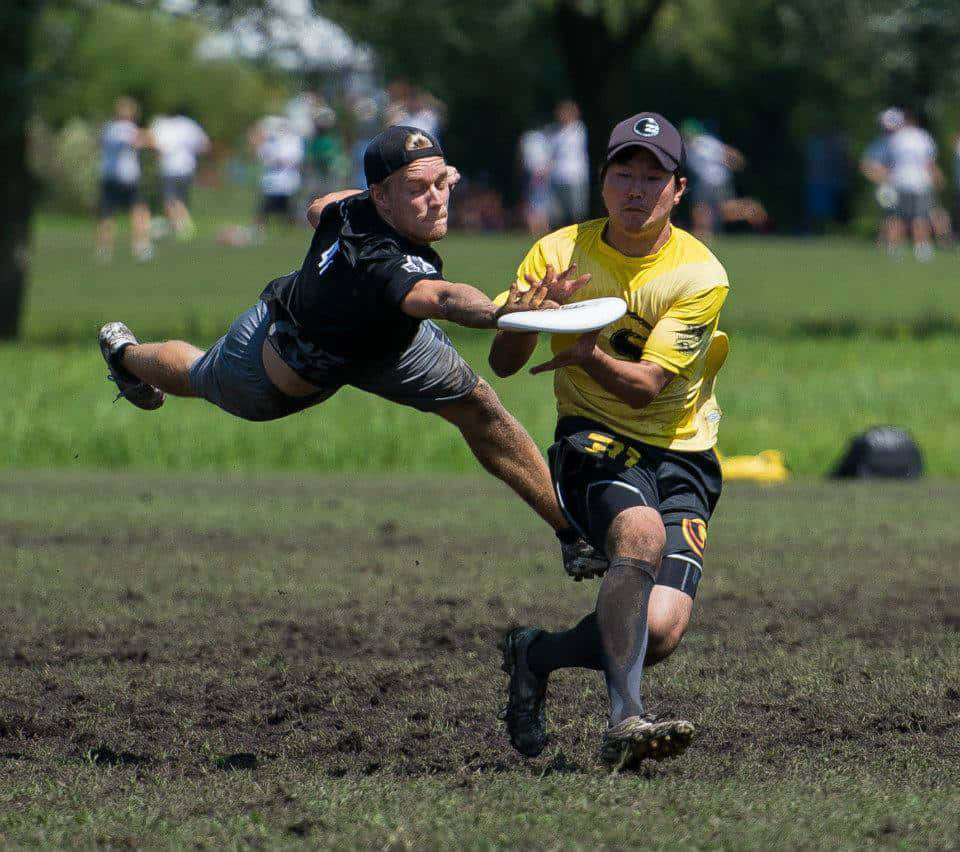 球員爭奪飛盤最佳 Ultimate Frisbee 背景 壁紙 — 兩名男性運動員在草地上激烈競爭，其中一人跳起攔截另一人飛來的飛盤。