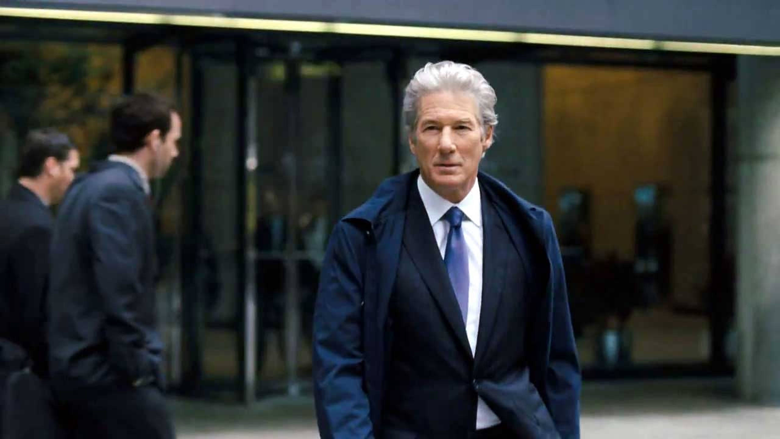 理查·基爾《 Arbitrage 》2012 壁紙 — 一位銀髮男士身穿深色西裝和藍色領帶，自信地走向鏡頭，背景是一座現代玻璃建築。