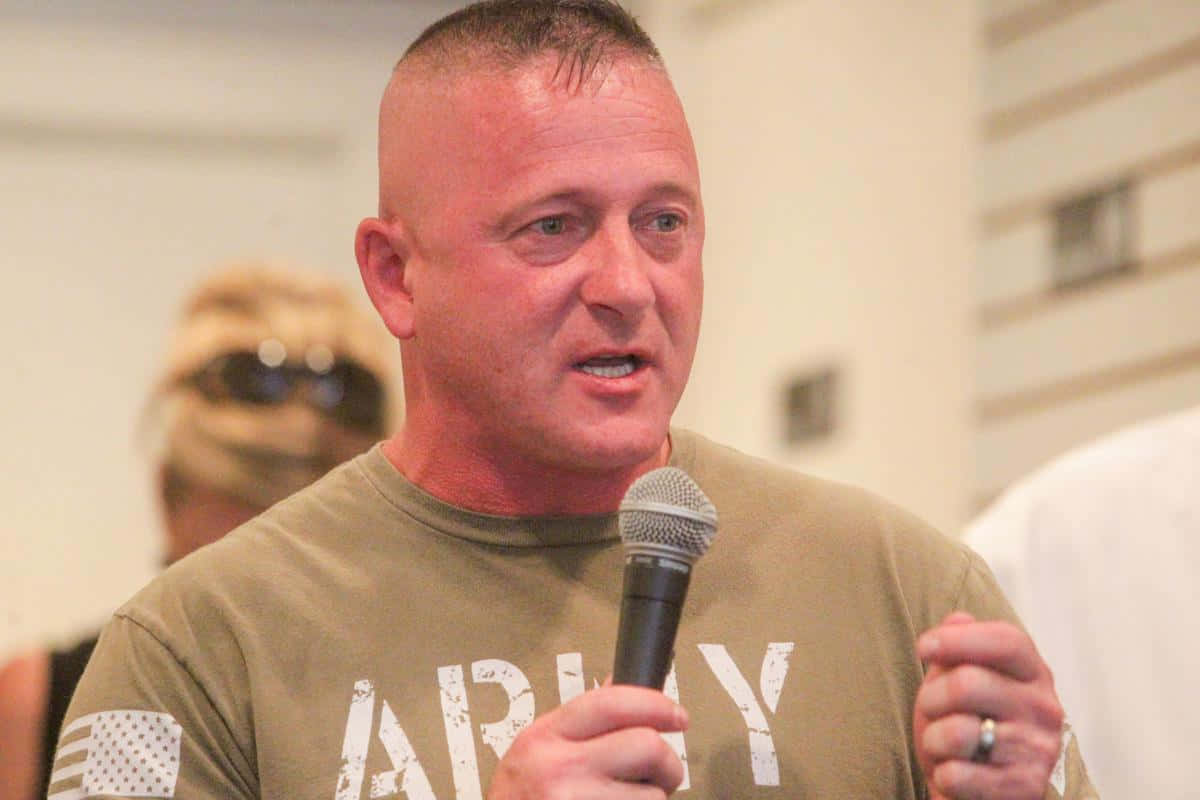 男子在室內對著麥克風說話，穿著軍裝T恤