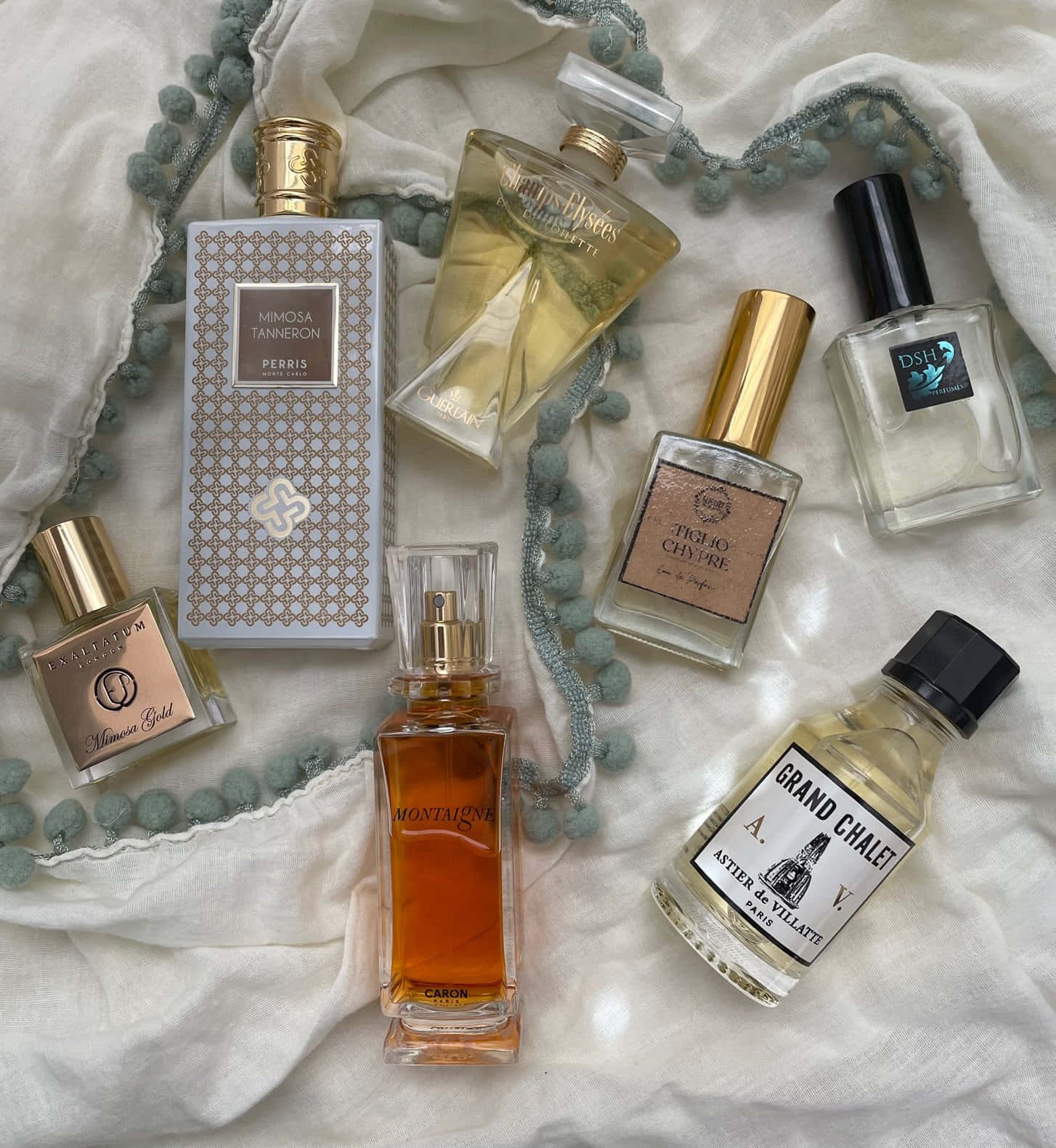 Spring Perfume 桌布 壁紙 — 琥珀色和透明玻璃的優雅香水瓶放在柔軟的白色布料上，配有藍綠色絨球飾邊。