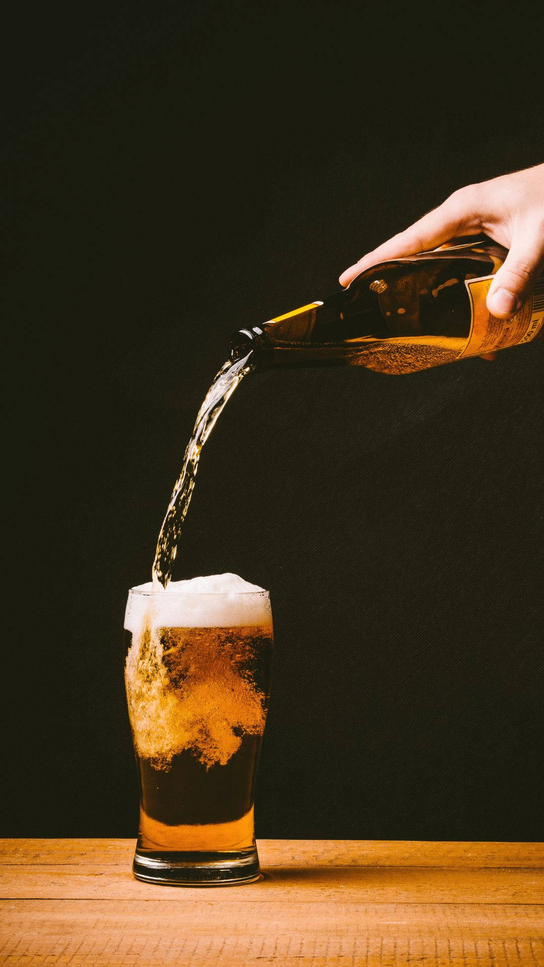 琥珀色酒精倒入杯中冒泡 壁紙 — 一隻手將金黃色啤酒倒入木桌上的玻璃杯中，背景為深色。