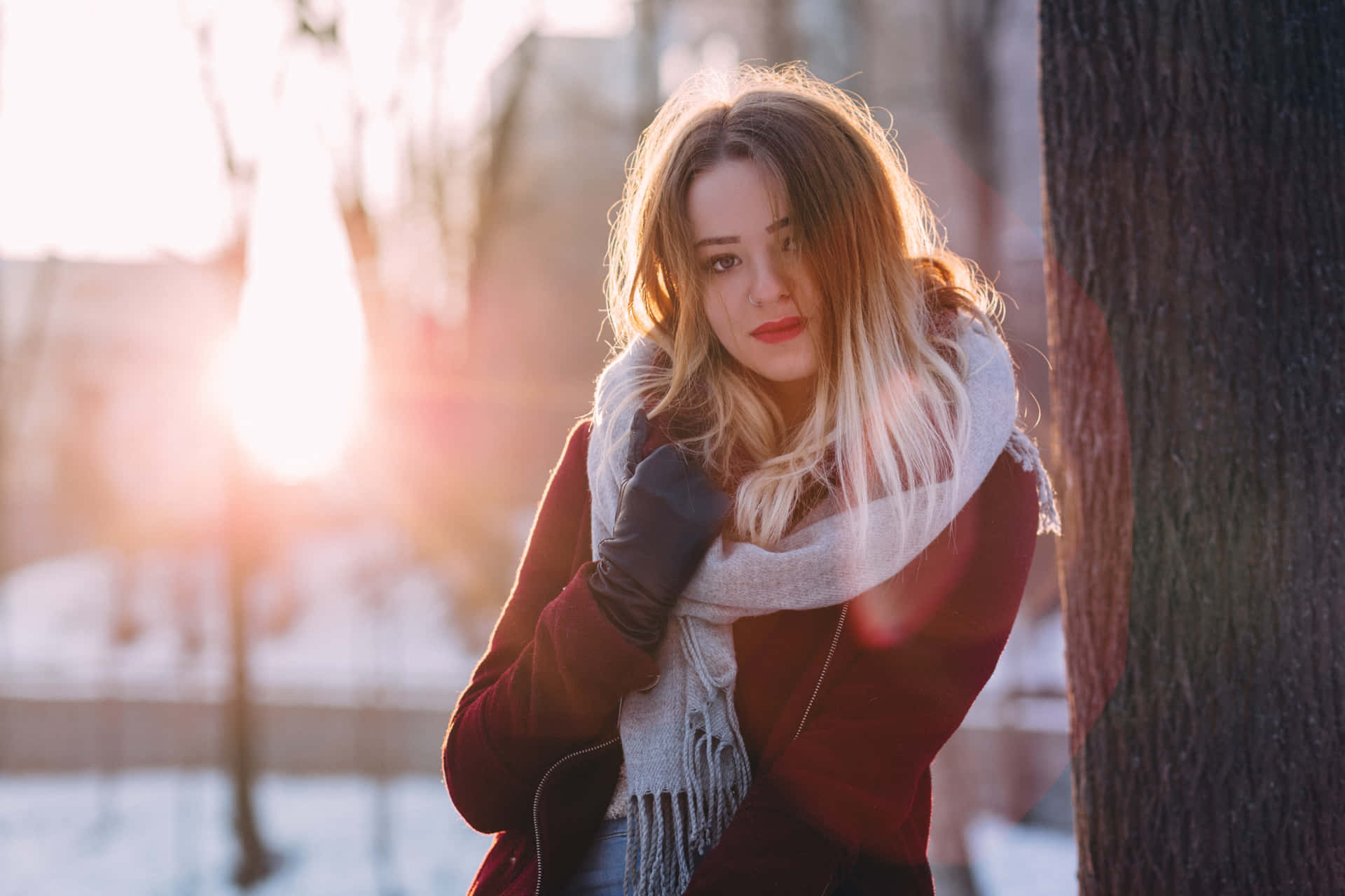 身穿冬季外套和圍巾的女性靠在雪地公園的樹上，陽光照射。