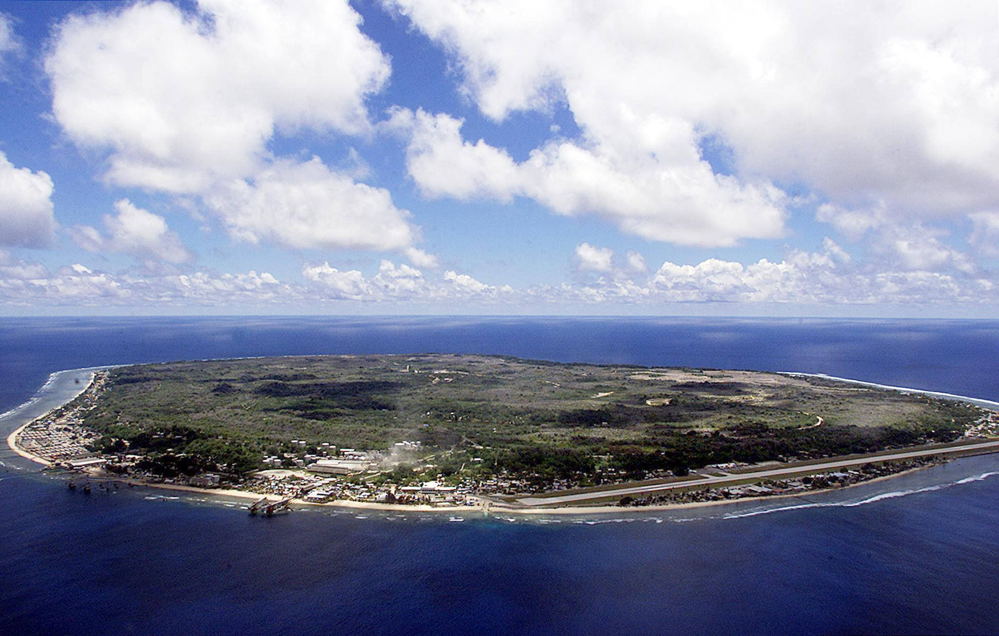 瑙魯島空拍全景 壁紙 — 從空中俯瞰一個鬱鬱蔥蔥的綠色島嶼，島上有沿海小鎮和蜿蜒的道路，被深藍色的海洋環繞，天空部分多雲。