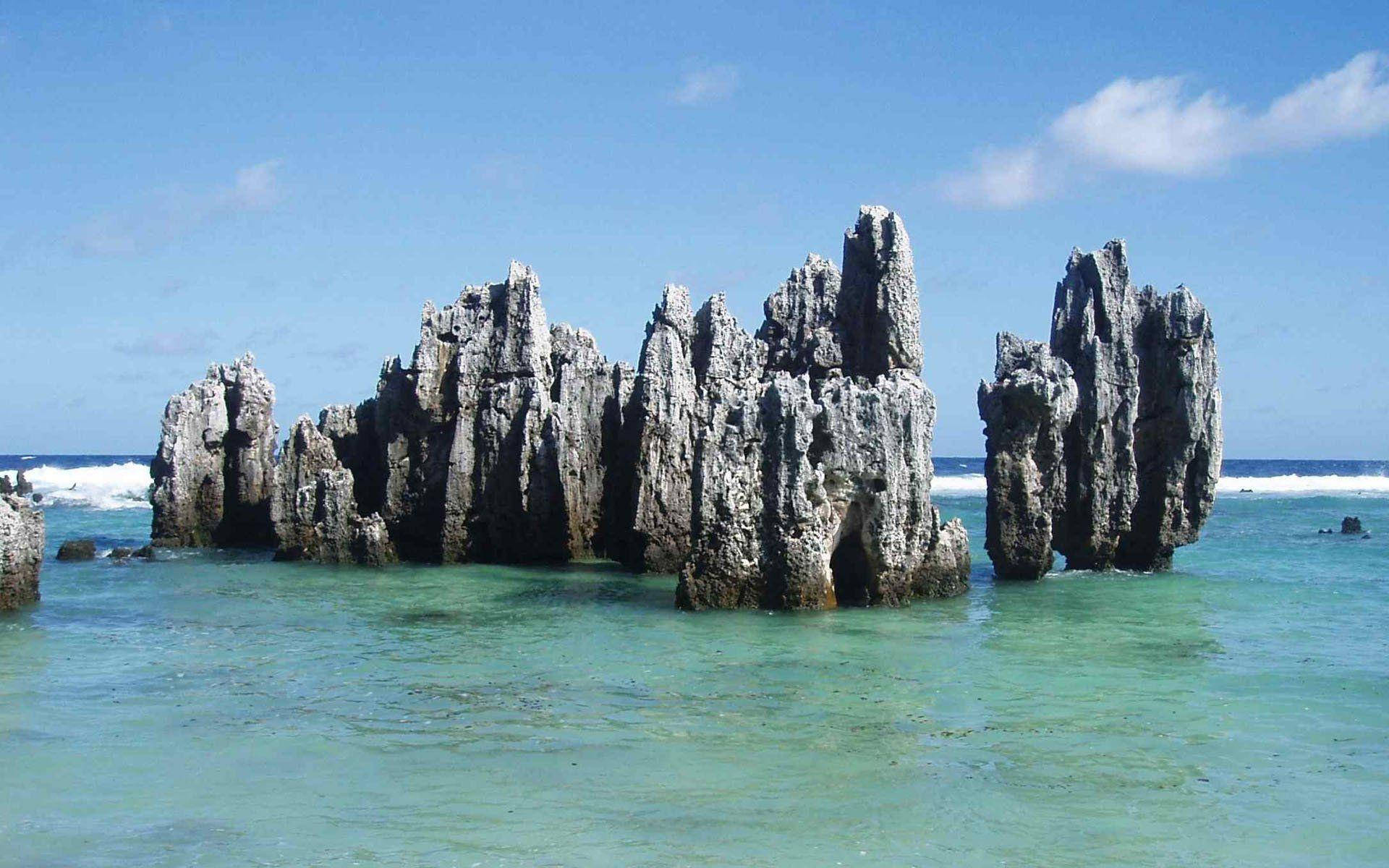 瑙魯阿尼巴雷灣的巨大岩層 壁紙 — 高聳的鋸齒狀岩石層從清澈的碧綠海水中升起，在藍天白雲下。