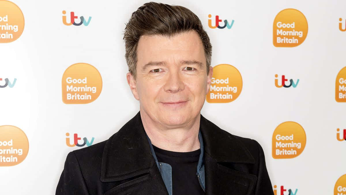 身穿黑色外套的男子在ITV Good Morning Britain背景的媒體活動中微笑。