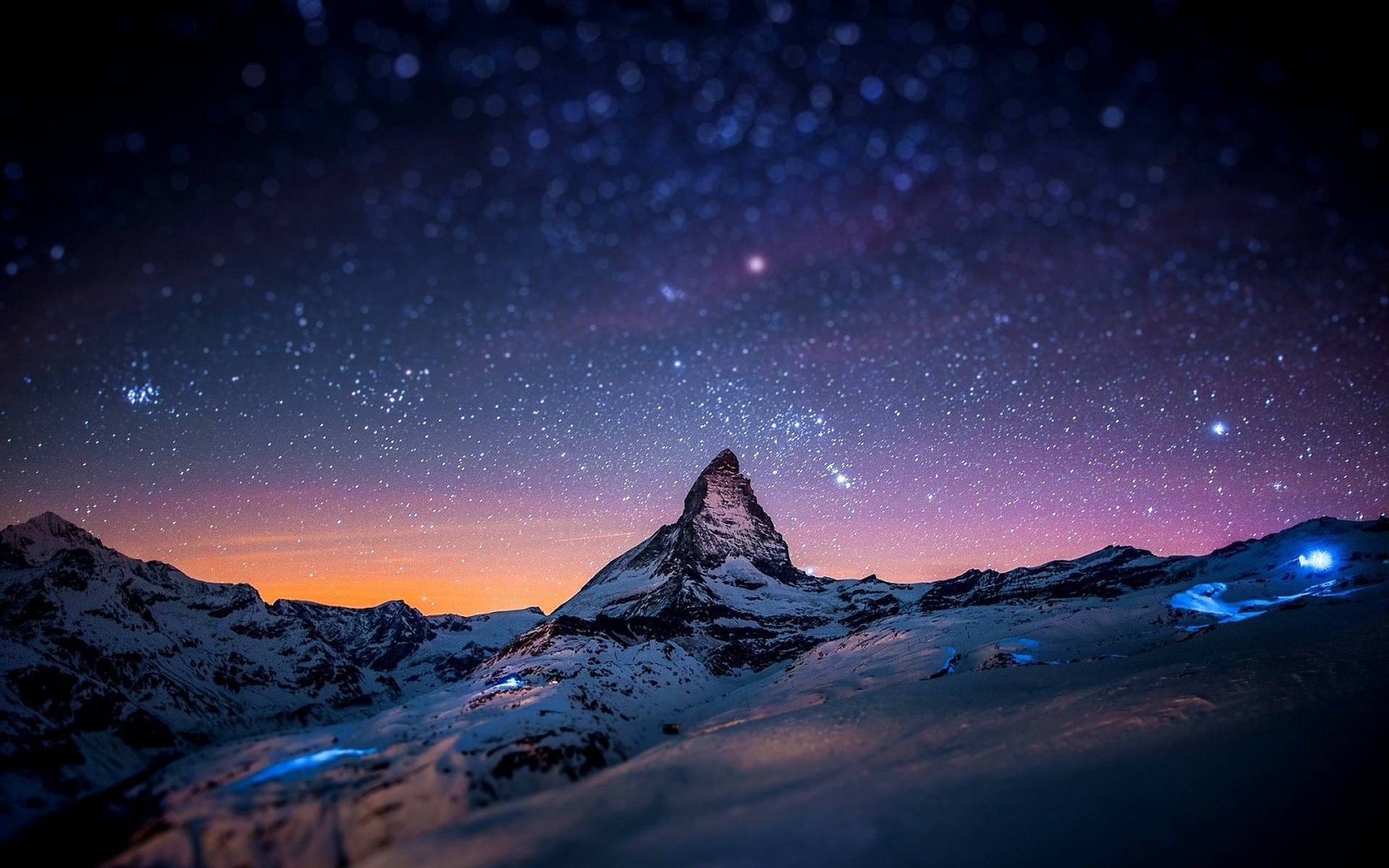 瑞士阿爾卑斯山夜景 壁紙 — 白雪覆蓋的馬特洪峰在繁星點點的暮色天空下聳立，地平線呈現紫橙色，下方有發光的藍色燈光。