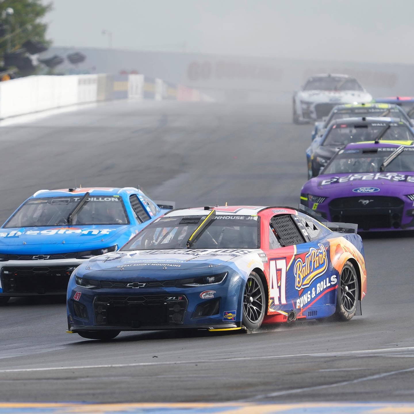 NASCAR賽車在潮濕的賽道上飛馳，背景有霧氣。