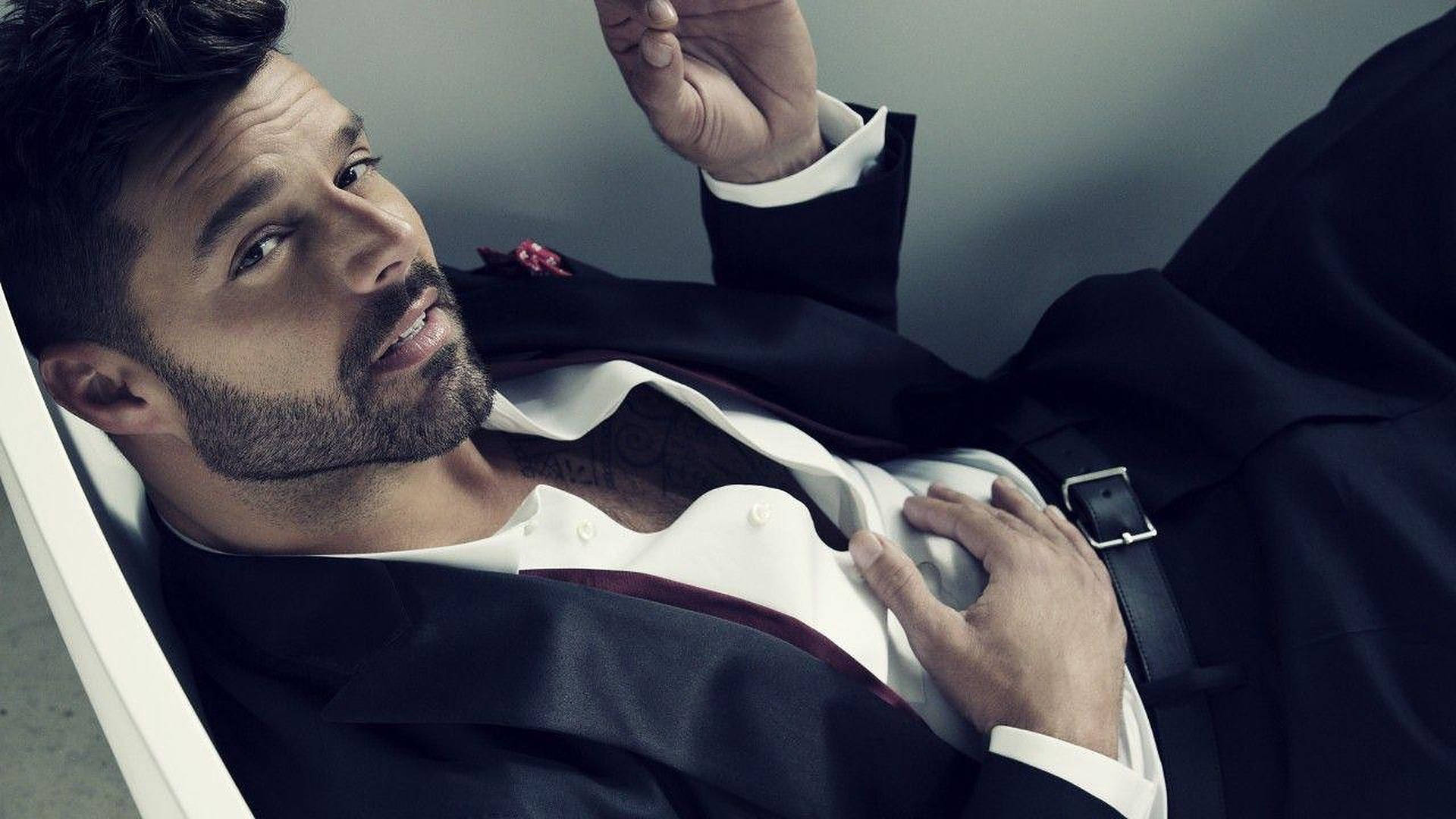 瑞奇·馬丁西裝照 壁紙 — 一位英俊男士身穿黑色西裝，隨意地靠著，眼神自信，佩戴紅色襟花。