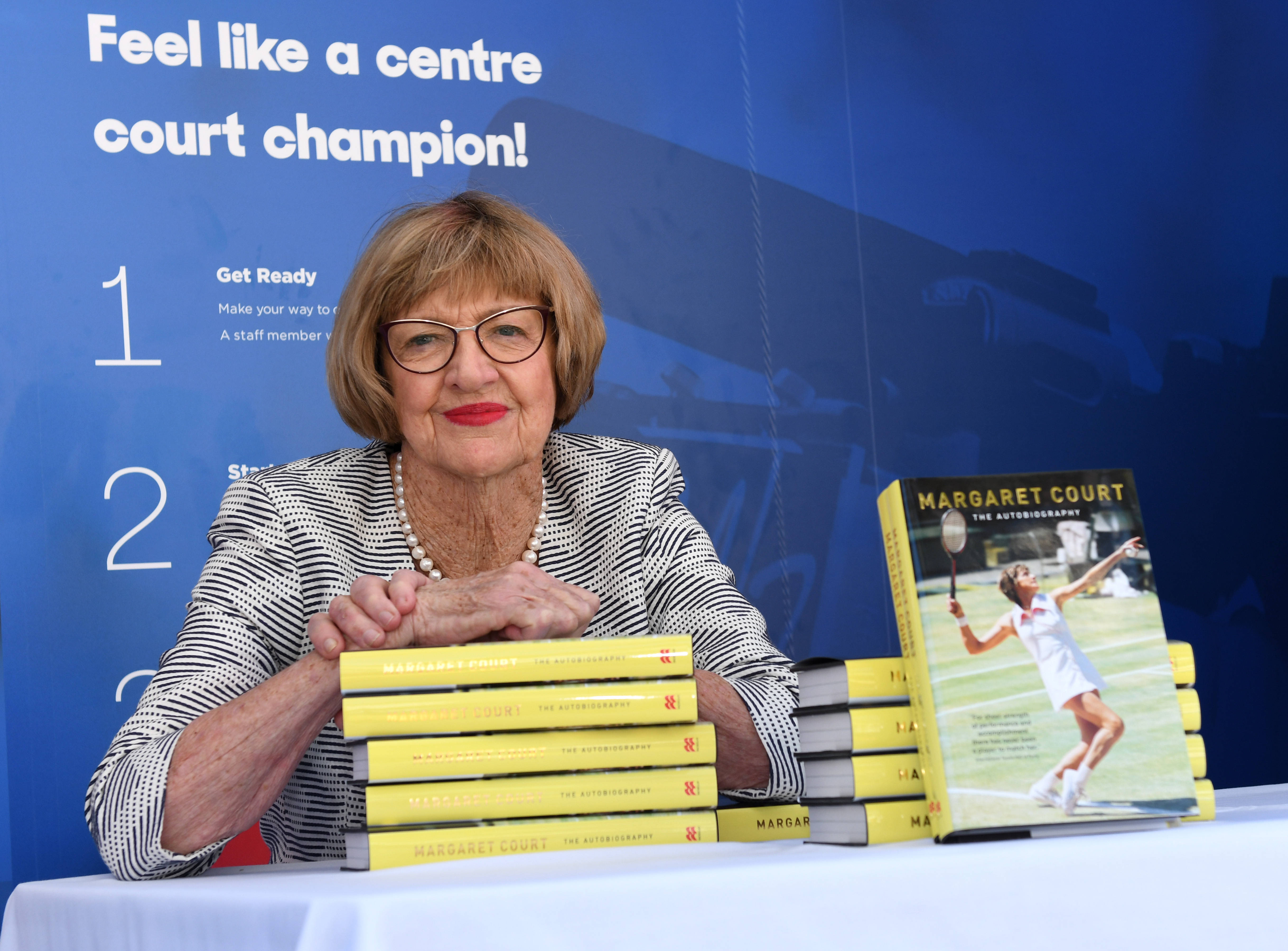 瑪格麗特·考特出席新書發表會 壁紙 — 年長嘅網球傳奇人物 Margaret Court 喺宣傳活動中坐喺一堆黃色自傳書後面，露出溫暖嘅笑容。