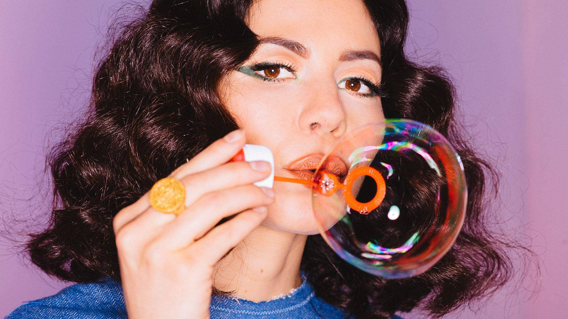 瑪莉娜與鑽石吹泡泡 壁紙 — 一位擁有深色捲髮的女性在柔和的紫色背景下吹出閃閃發光的彩虹泡泡。
