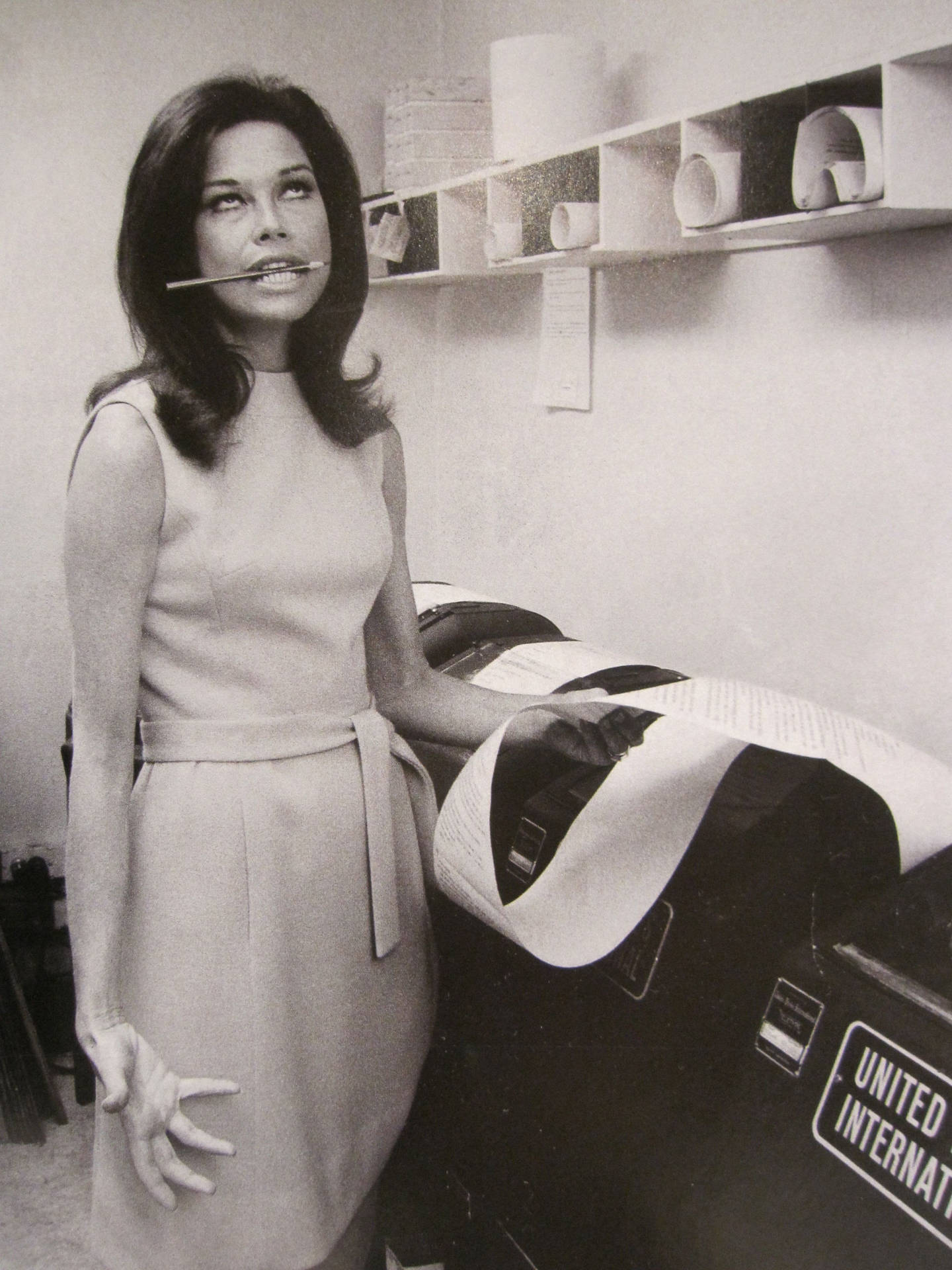 身穿1960年代連衣裙的女性，在辦公室郵件機旁嘴裡叼著筆。