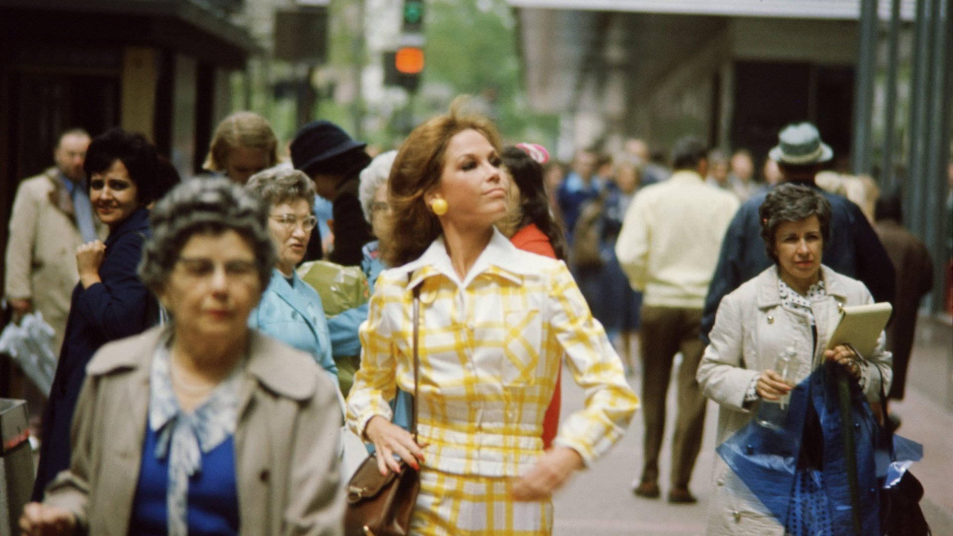 身穿黃色格子西裝的女性走過1970年代繁忙的城市街道。