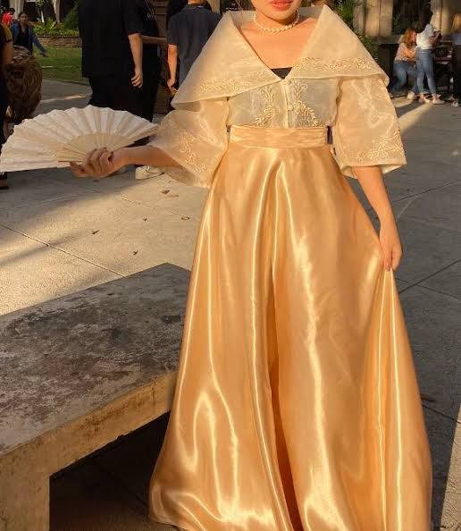 身穿金色傳統禮服的女性在日落時分於戶外手持白色扇子。