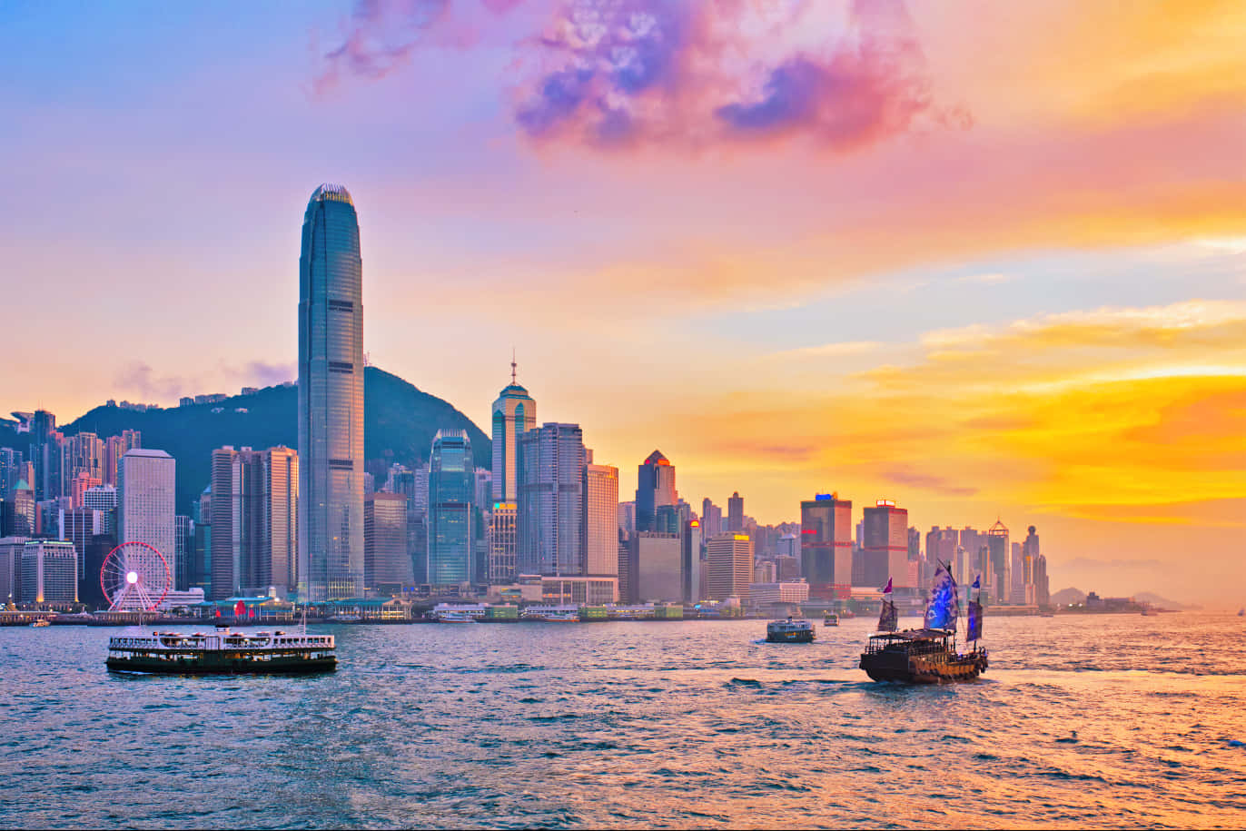 璀璨奪目的香港天際線夜景 壁紙 — 香港日落時分嘅天際線，充滿活力嘅橙色雲彩倒映喺海港水面，船隻喺高聳嘅摩天大廈旁邊滑過。