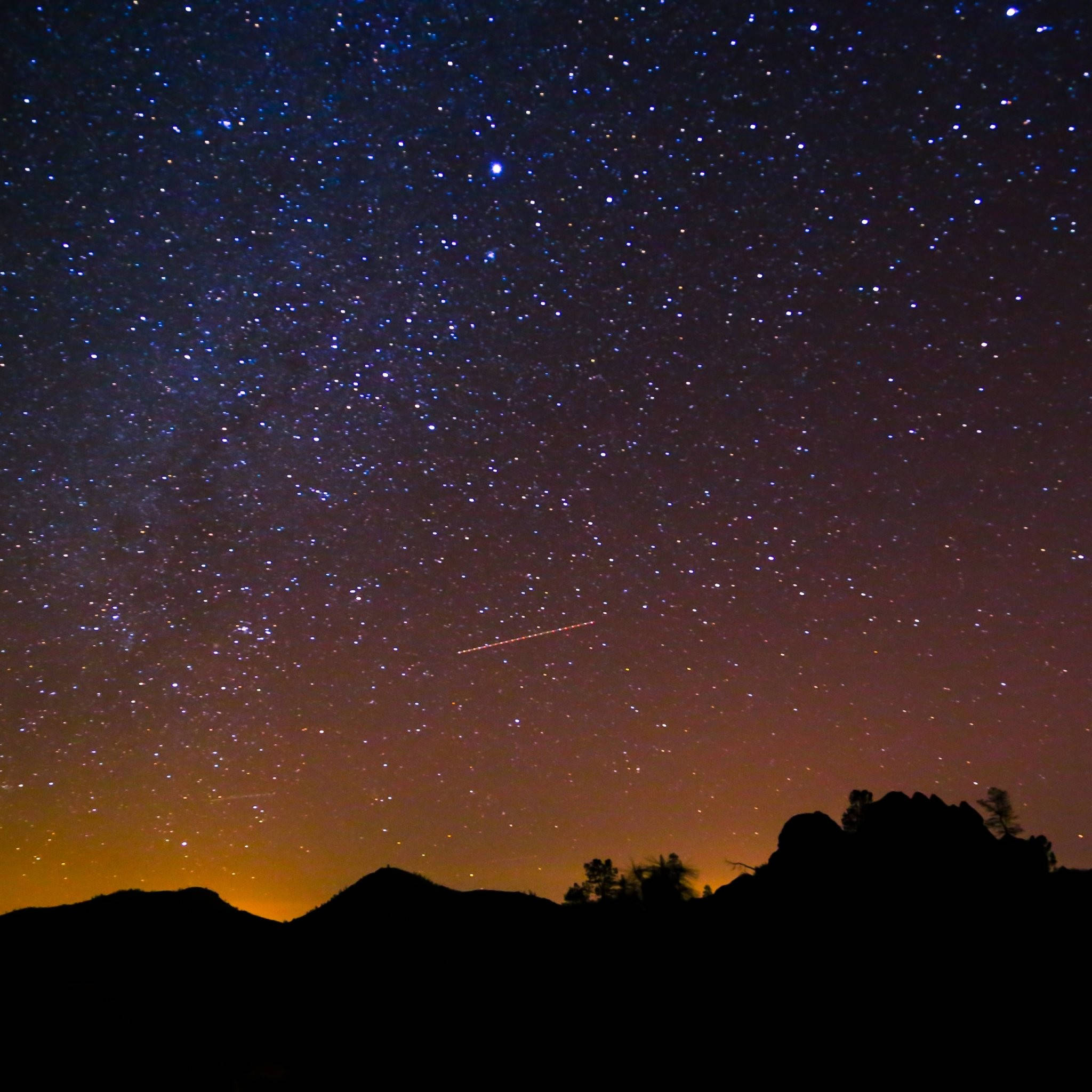 璀璨星河與銀河夜空高清壁紙 壁紙 — 繁星點點的夜空，一顆明亮的流星劃過黑暗的山脈輪廓，映襯著橙色的地平線。