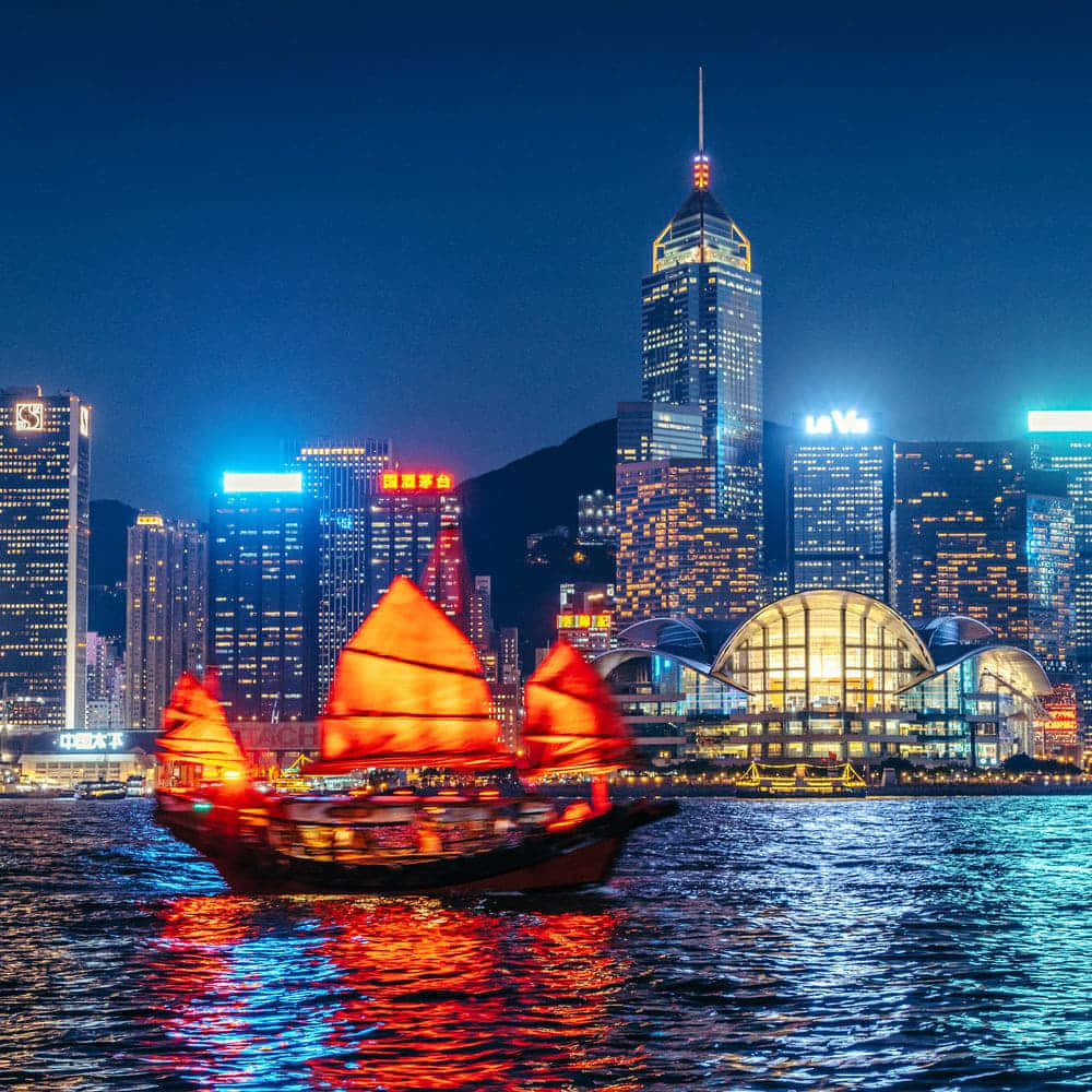 璀璨香港夜景天際線 壁紙 — 一艘傳統中式帆船，帶有發光的紅色船帆，在夜間滑過港口，反射著高聳摩天大樓的霓虹燈光。