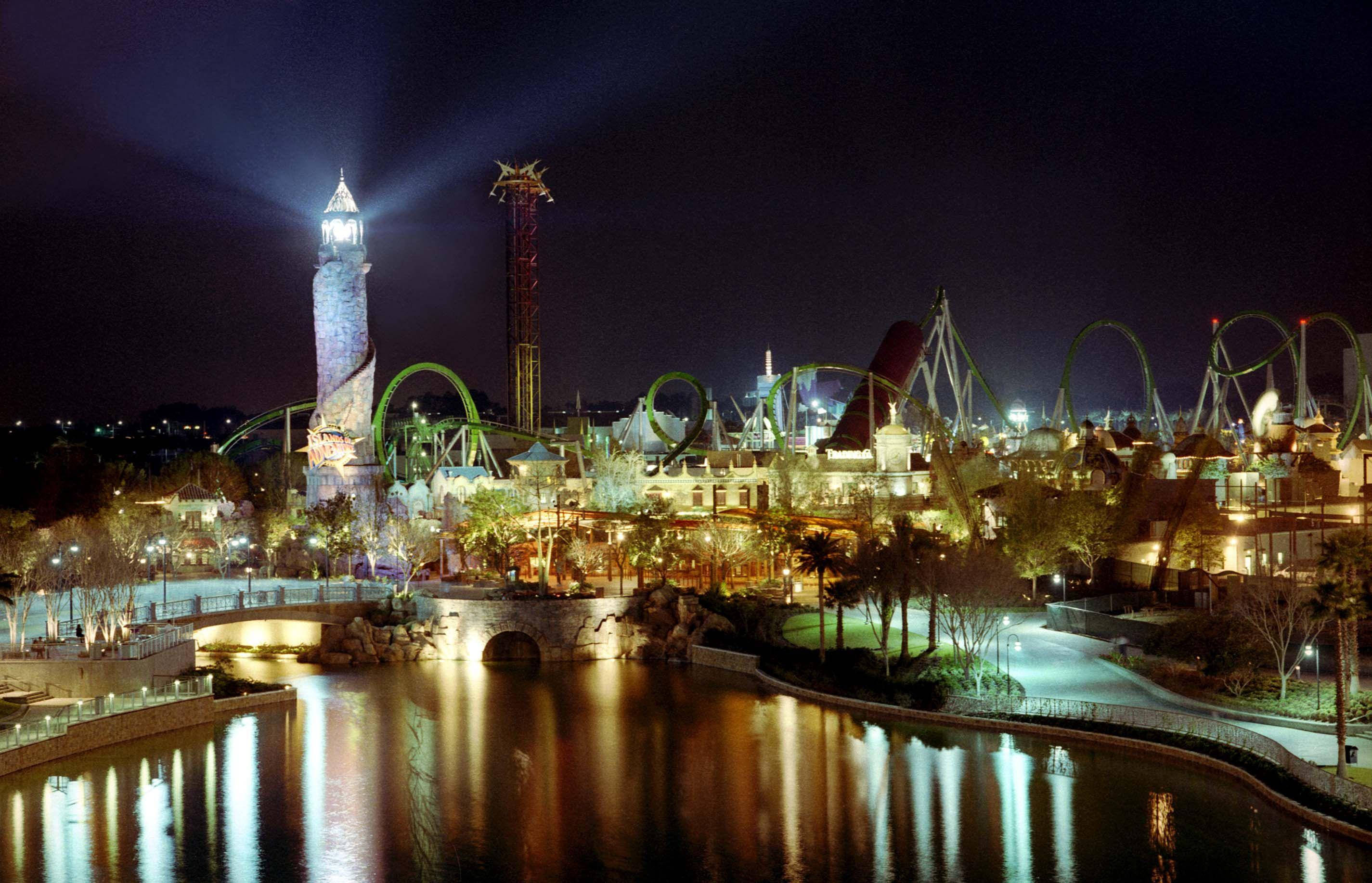 環球影城夜間鳥瞰全景 壁紙 — 一個耀眼的夜間遊樂園，發光的過山車和點亮的塔樓倒映在平靜的水道上。