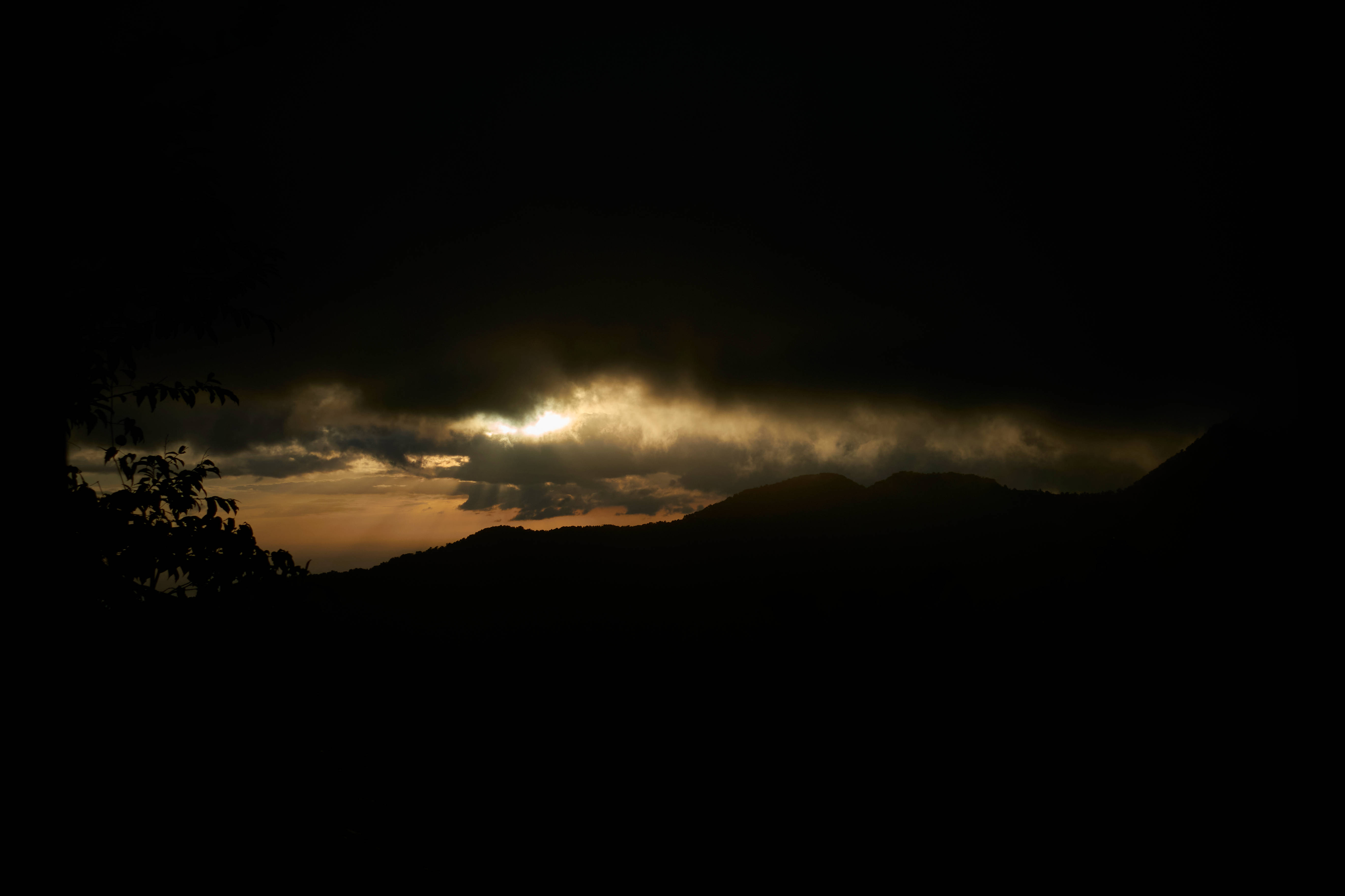 瓜德羅普夜空 壁紙 — 戲劇性的金色陽光穿透黑暗的暴風雲，照耀在輪廓分明的山脈和樹枝上。